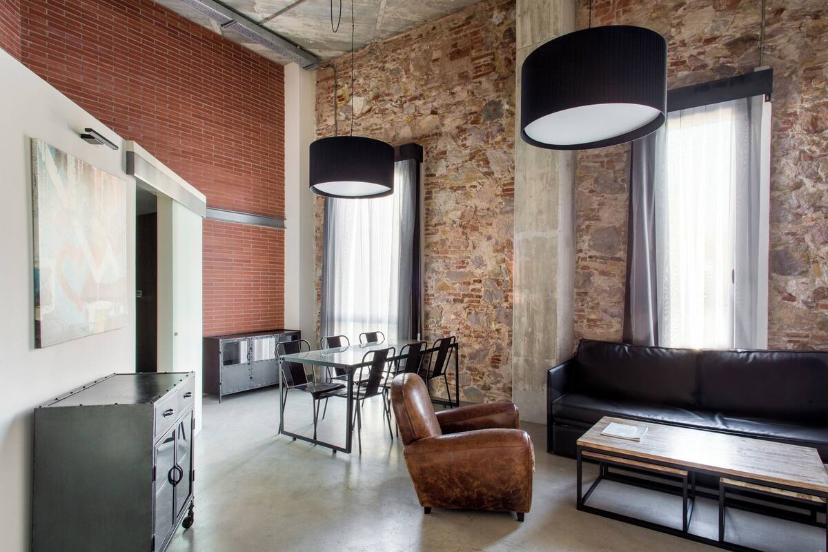 A modern interior with a brick wall, a black leather sofa, a dining table with chairs, and pendant lighting.