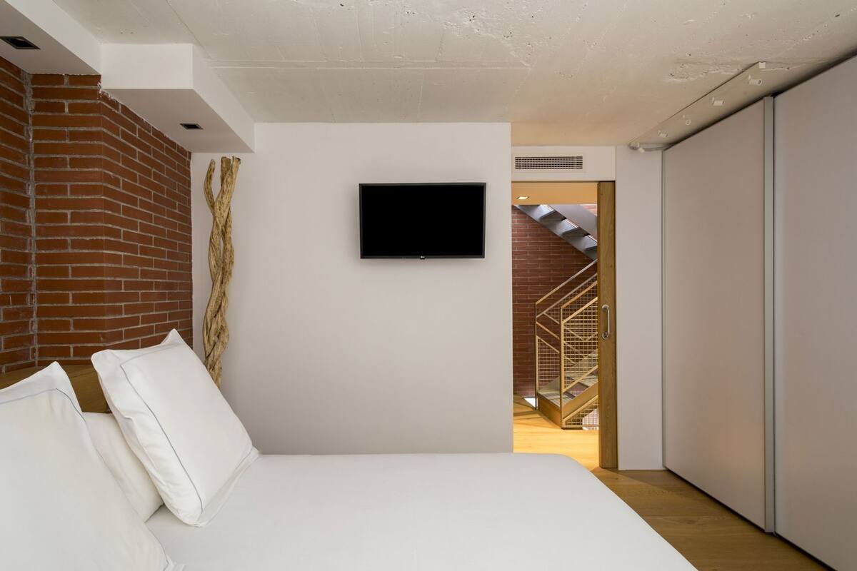A bedroom with a bed, a TV mounted on the wall, a brick wall, and a staircase leading to another room.
