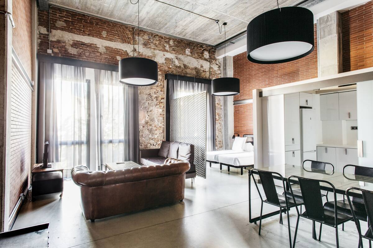 A modern interior with a leather sofa, dining table, and chairs, a brick wall, and large windows with curtains.