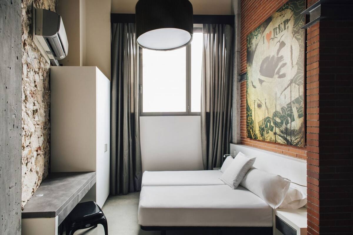 A hotel room with a bed, a desk, a chair, a window with curtains, a hanging light fixture, and a wall mural.