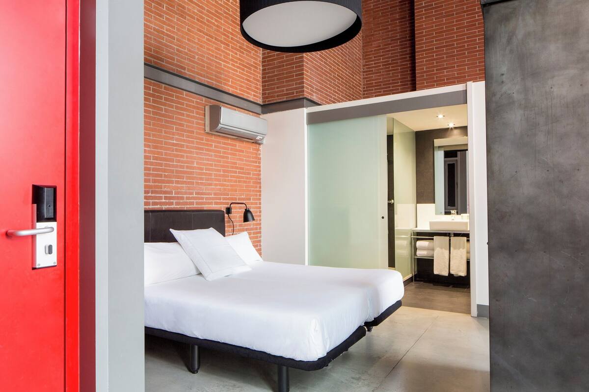 A modern hotel room with a brick wall, a bed with white linens, a red door, and a visible bathroom with a sink and towel rack.
