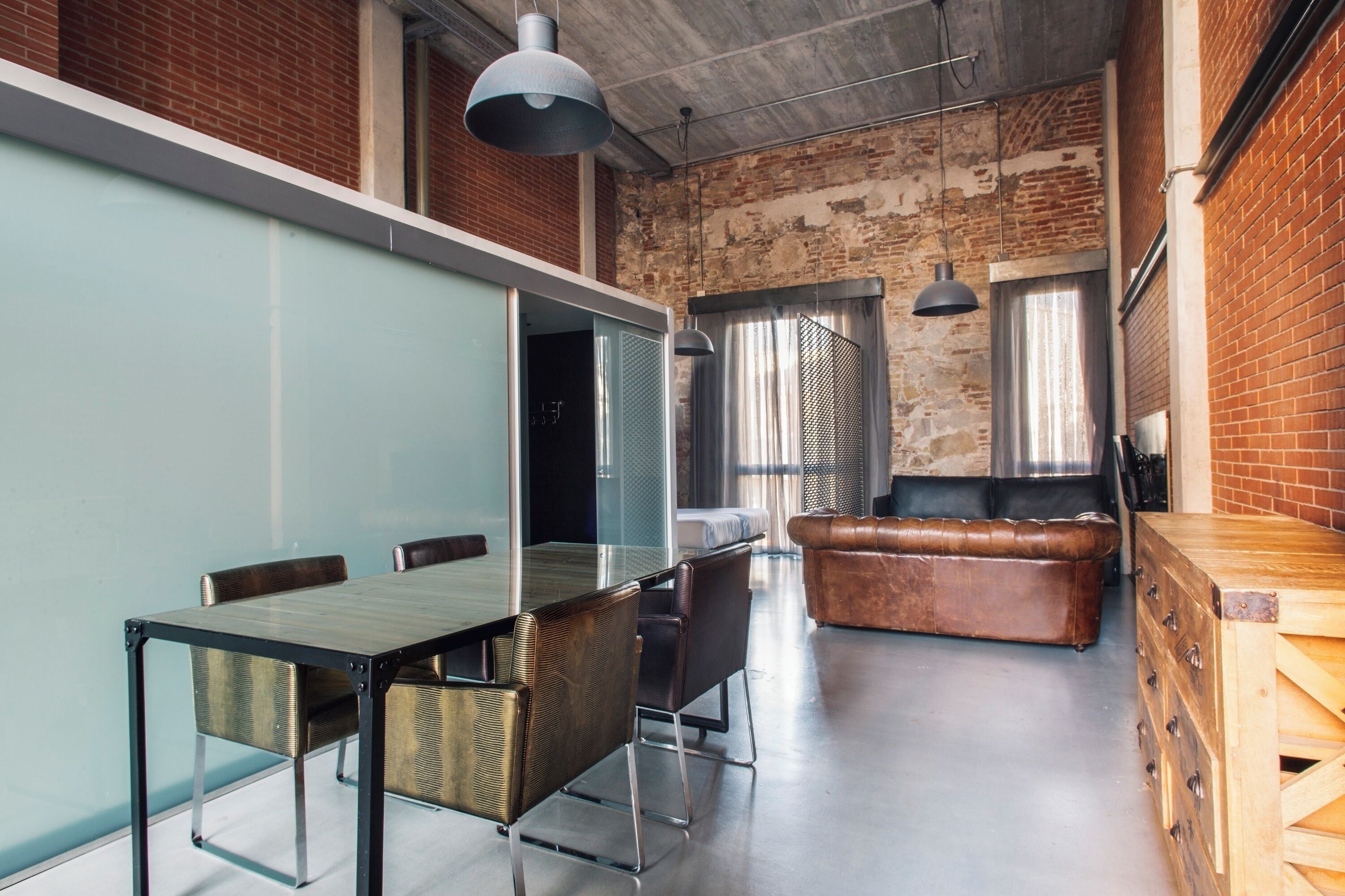 A modern office space with a glass partition, a wooden conference table, leather seating, and hanging pendant lights.
