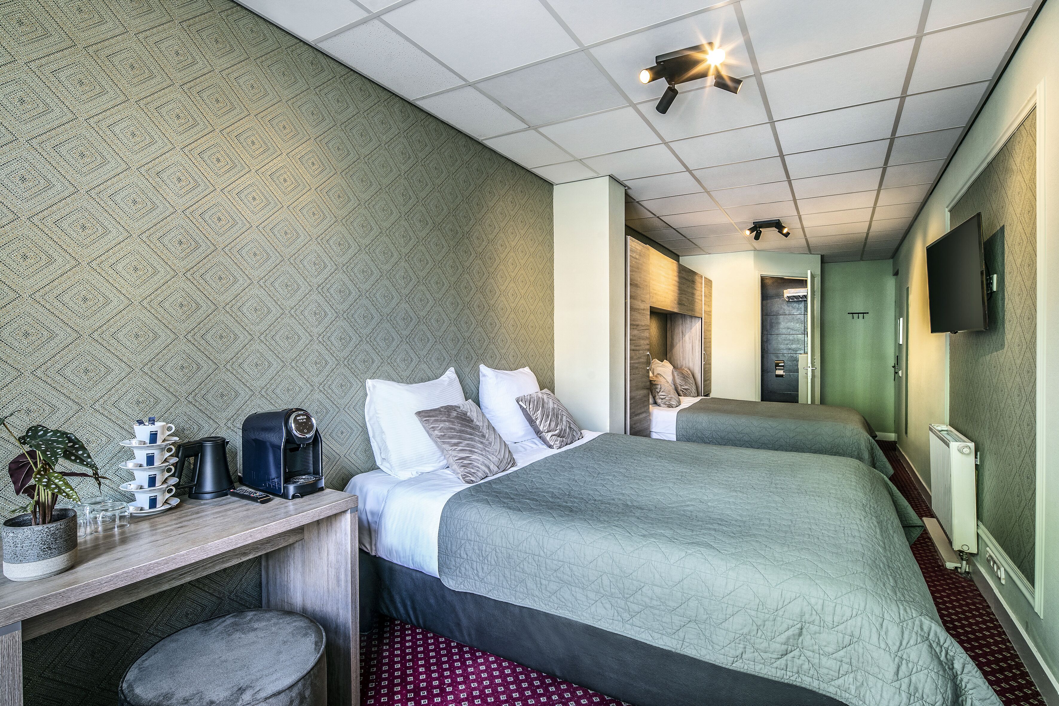 A hotel room with two beds, a desk with a coffee maker, a plant, and a TV mounted on the wall.