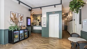 Un vestíbulo de hotel con piso de madera en patrón de espiga, una recepción, un refrigerador con bebidas y una planta colgante.