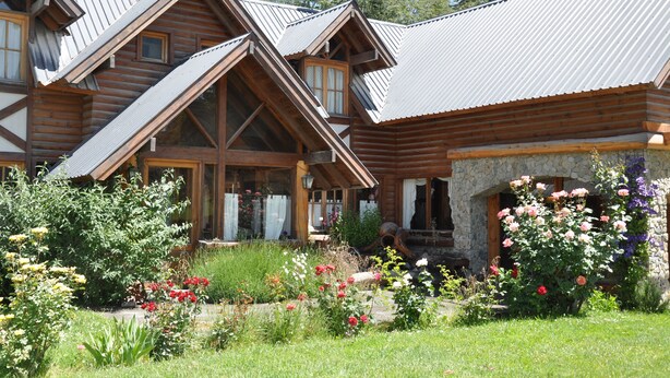 A wooden log cabin with a stone foundation, large windows, and a garden with various flowers. - Portal de Piedra (Villa La Angostura)