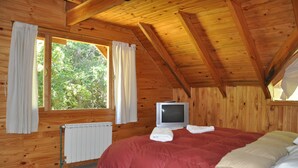 A wooden cabin bedroom with a bed, a television, and a window with curtains. - Portal de Piedra (Villa La Angostura)