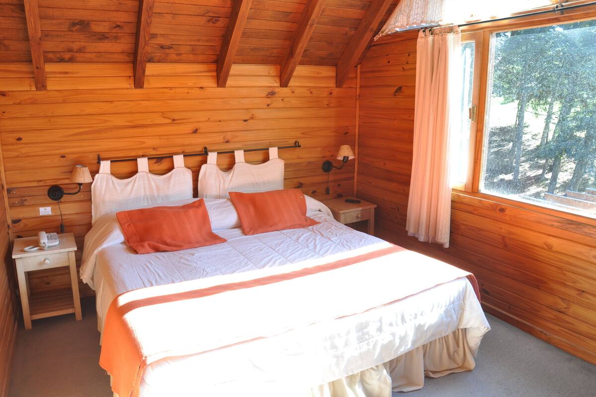 A wooden cabin bedroom with a bed, nightstands, and a window with curtains.