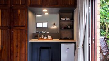 A compact kitchen with wooden cabinets, a small refrigerator, a bar stool, and a window overlooking greenery.
