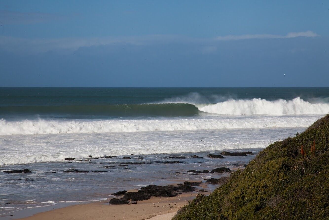 Una playa con olas rompiendo en la orilla, arena y un promontorio rocoso con vegetación.