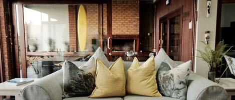 A cozy seating area with a grey sofa adorned with various cushions, a brick fireplace, and a surfboard mounted on the wall.