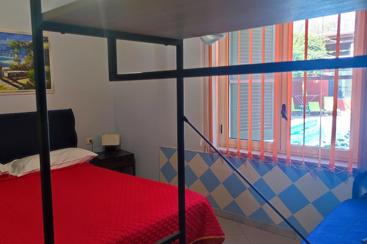 A bedroom with a red bed, a nightstand, a painting on the wall, and a view of the outside through a window with orange blinds.