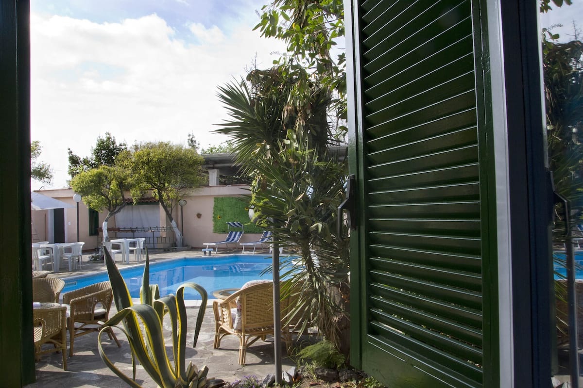 A pool area with lounge chairs and a small table, surrounded by plants and a green shutter.