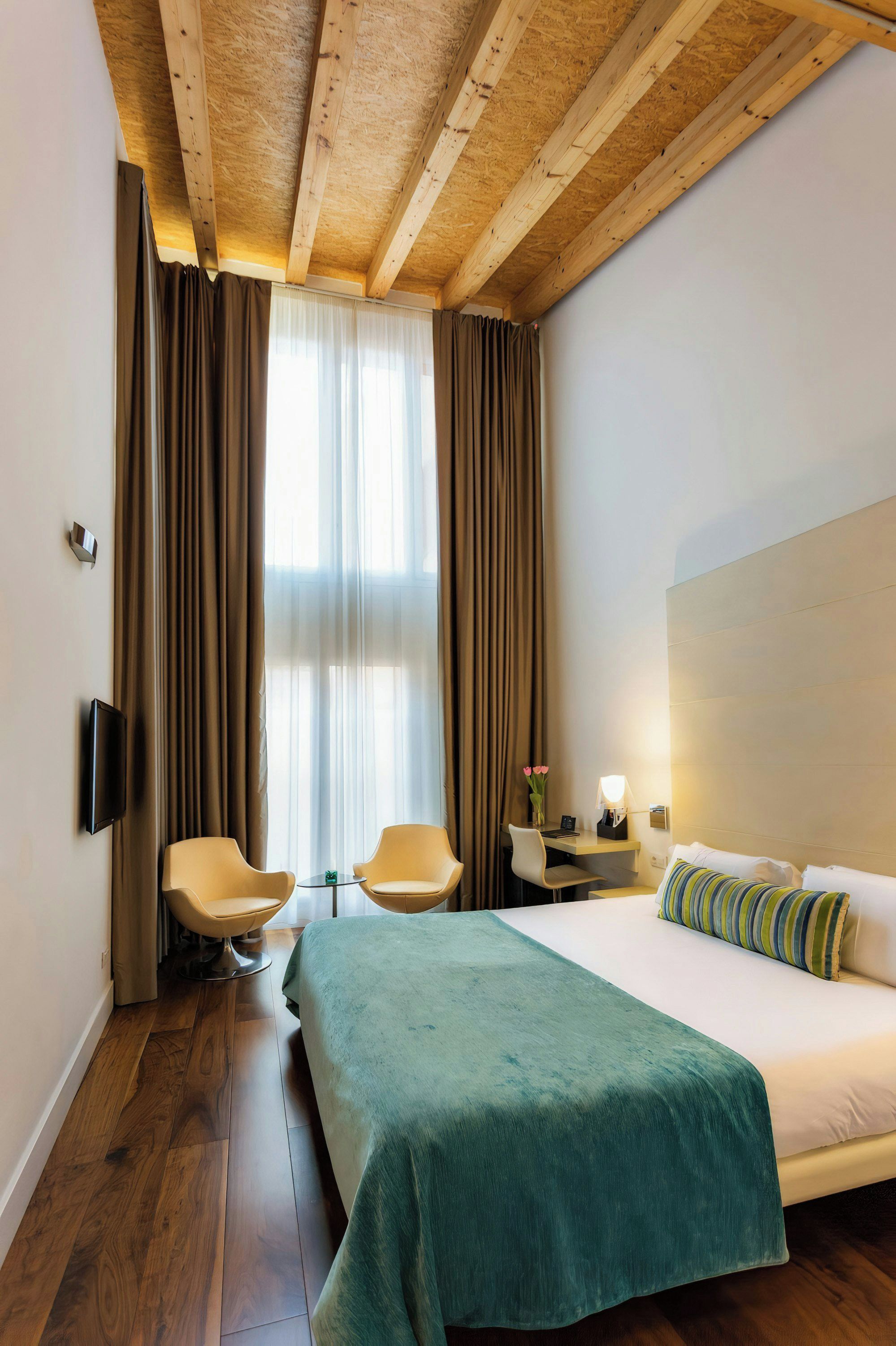A hotel room with a bed, two chairs, a desk, a TV, and a window with curtains.