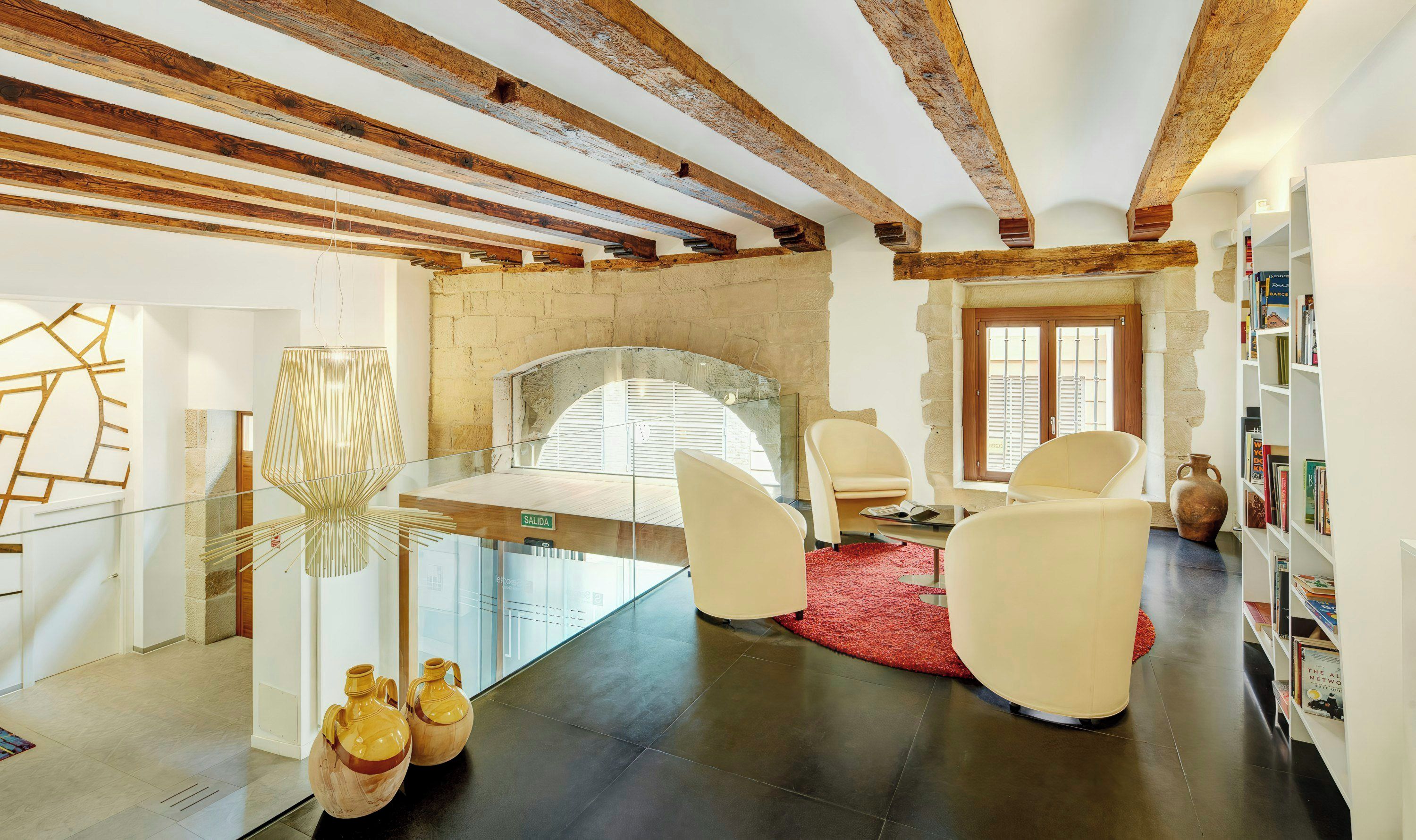 A modern interior with exposed wooden beams, a glass table, white chairs, a red rug, and a bookshelf.