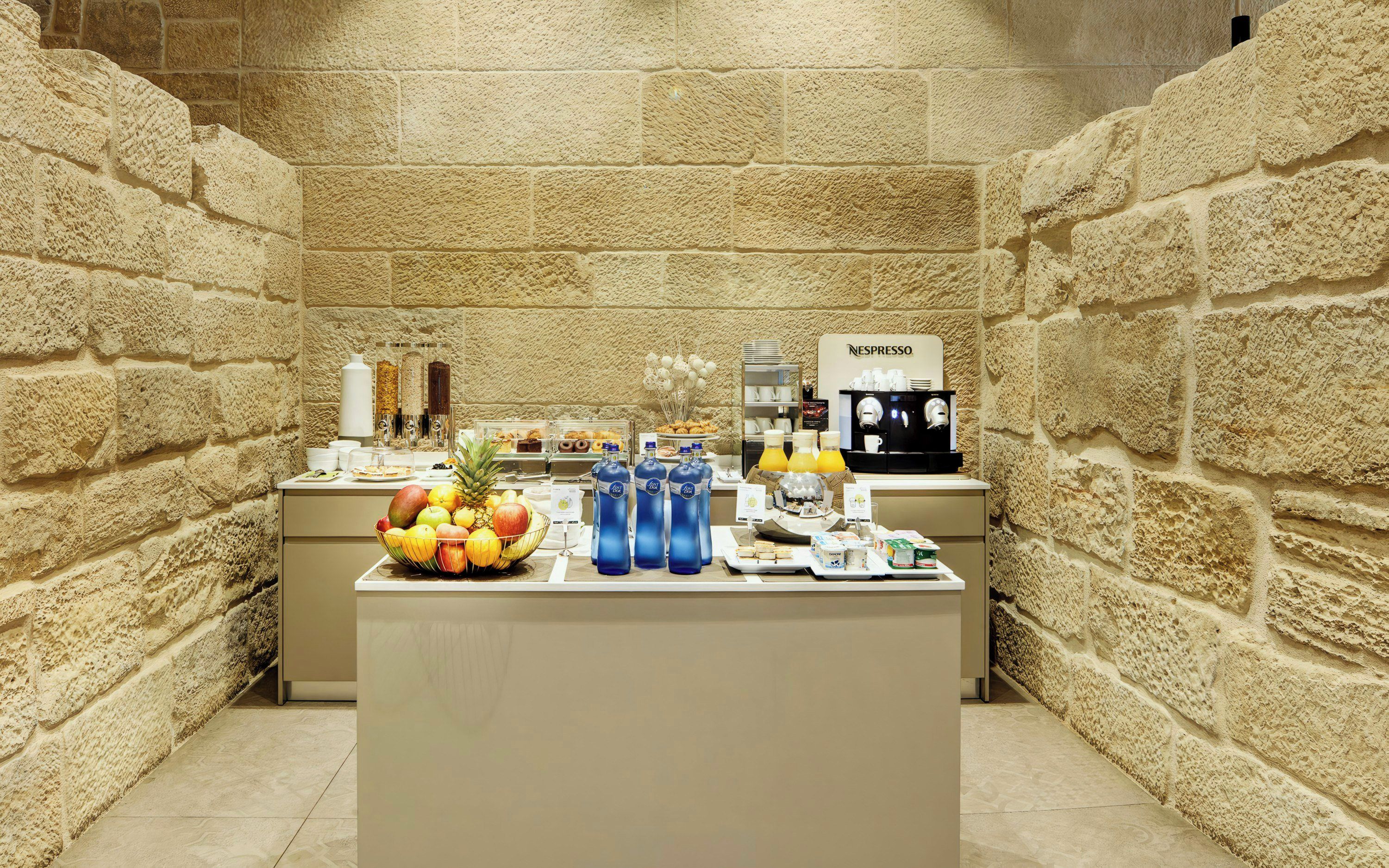 Un bufé de desayuno con un fondo de pared de piedra, que incluye frutas, bebidas y una máquina de café.
