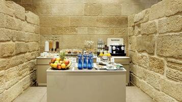 Un bufé de desayuno con un fondo de pared de piedra, que incluye frutas, bebidas y una máquina de café.