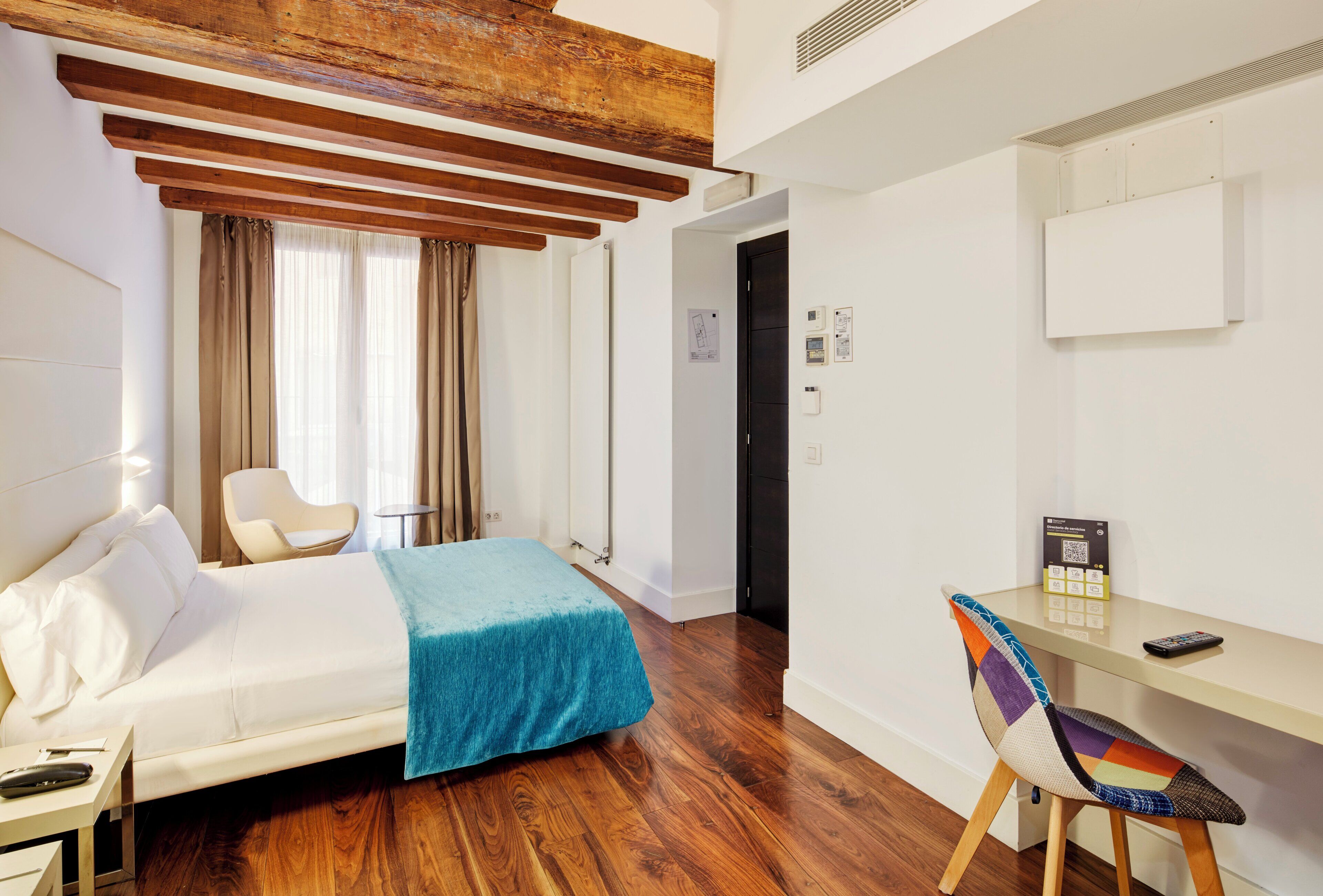 A hotel room with a bed, a desk, a chair, and a wooden ceiling.