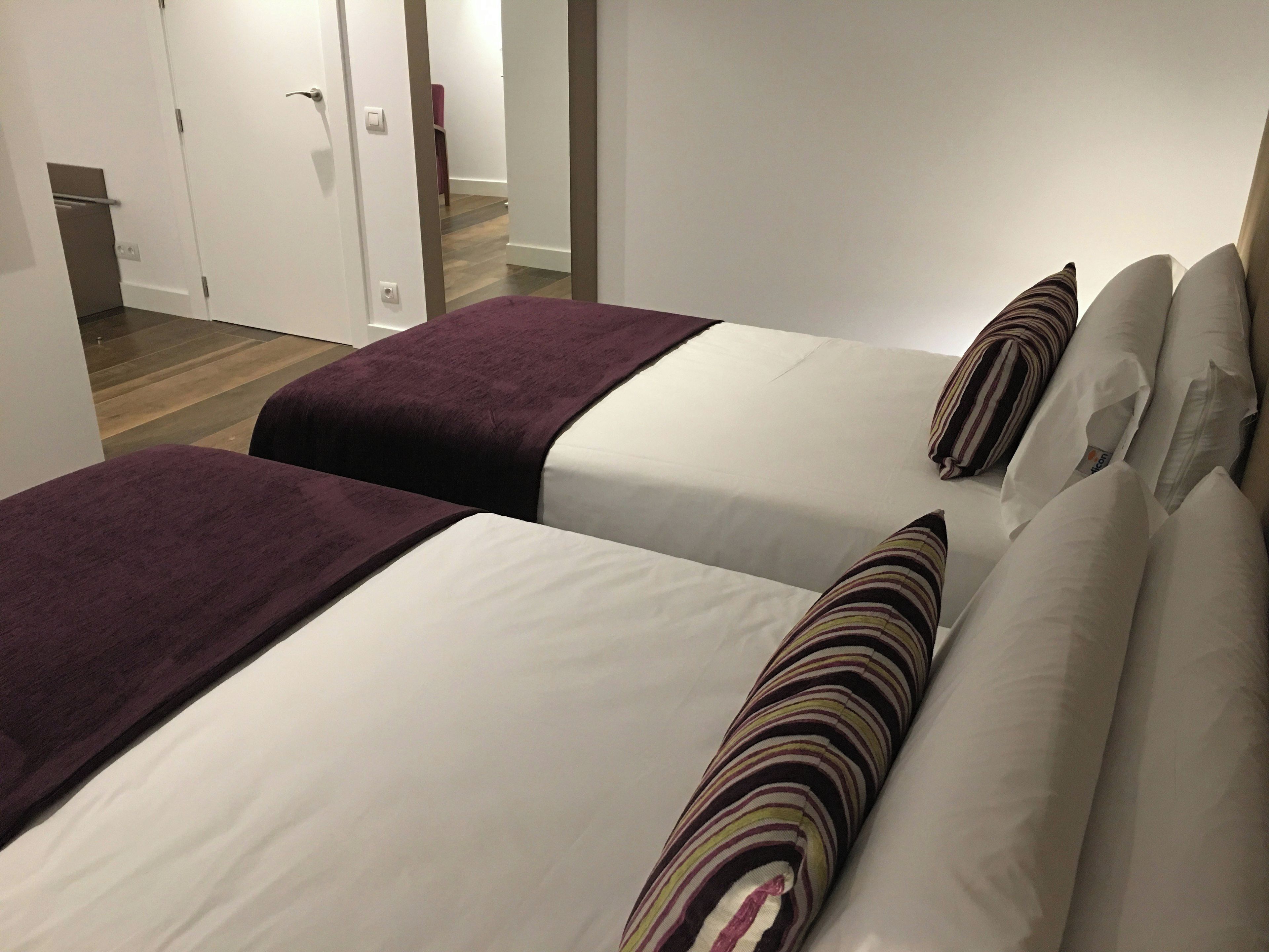 Une chambre d’hôtel avec deux lits, chacun doté d’un matelas blanc et d’une couverture violette. Des oreillers à rayures sont posés sur les lits.