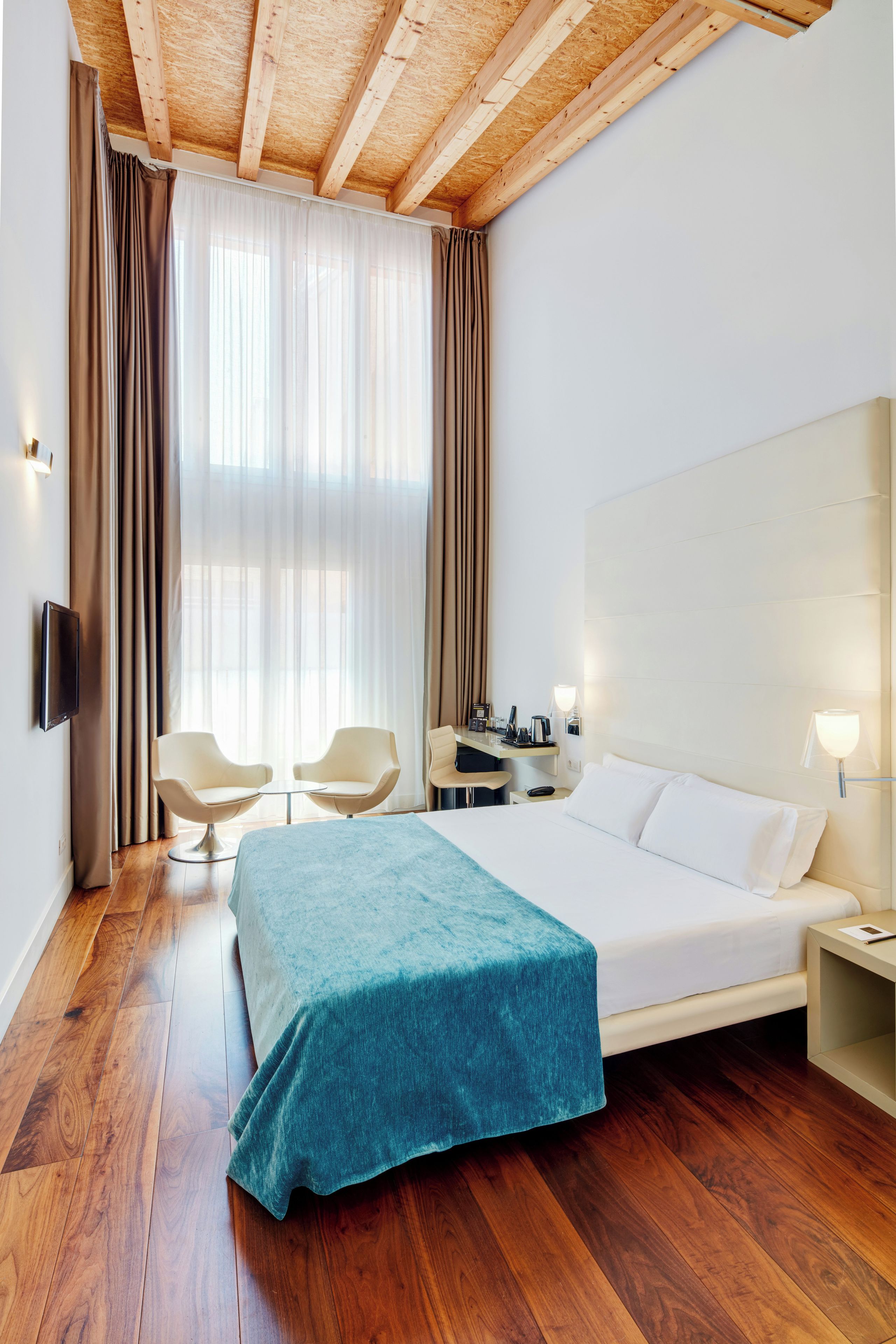 Een hotelkamer met een bed, een bureau, twee stoelen, een televisie en een groot raam met gordijnen.