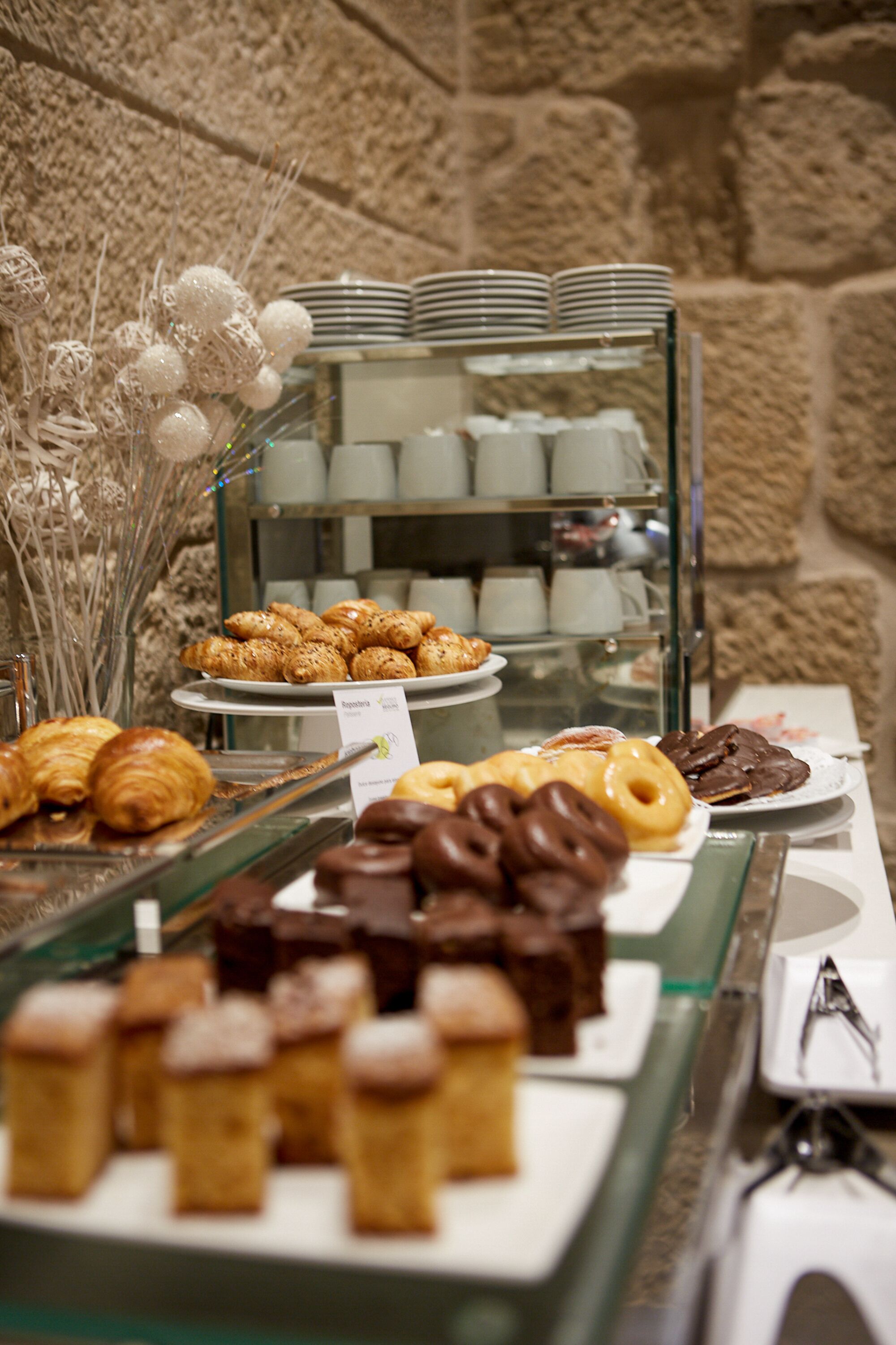 Un bufé con diversos pasteles y productos de panadería, incluyendo croissants y rosquillas, expuestos en un escaparate de vidrio.