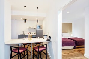 A modern kitchen with a microwave, sink, and bar stools. There are two beds with red bedding in the adjacent room.