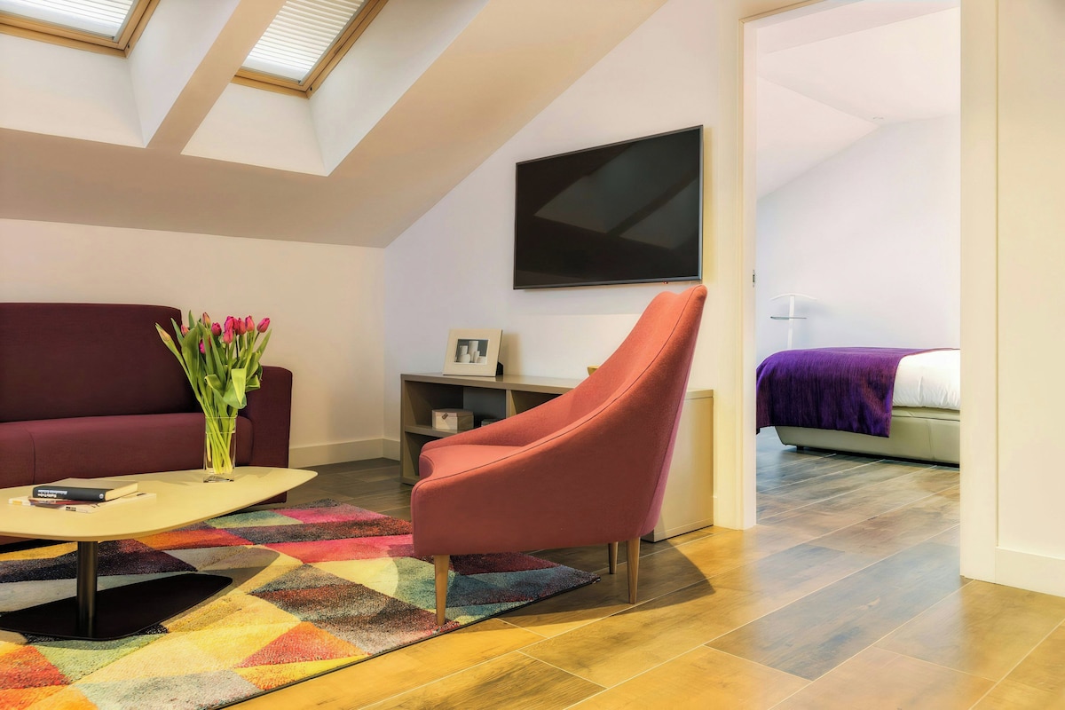 A modern living room with a sofa, a red chair, a flat-screen TV, and a bed with a purple blanket.
