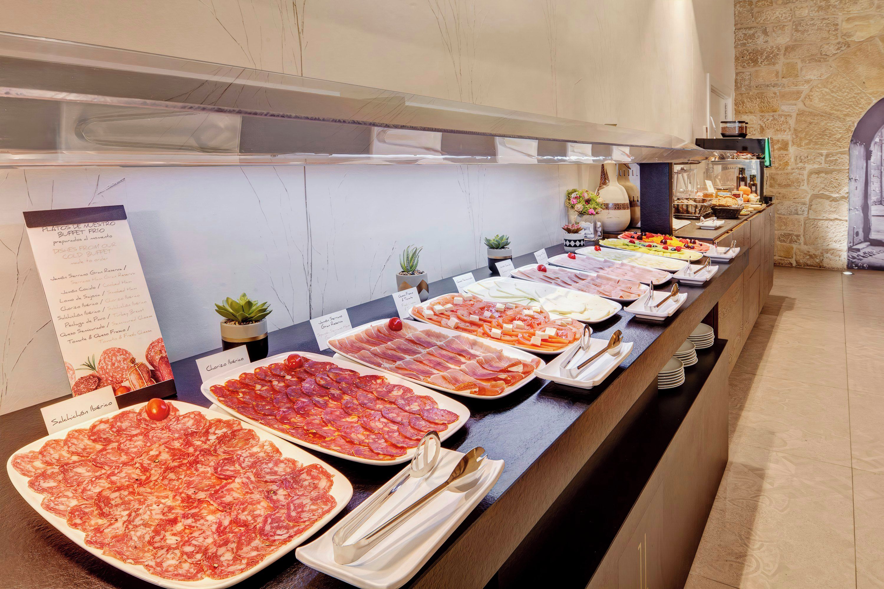 Un bufé con diversos platos fríos y carnes, incluyendo chorizo ibérico y salchichón ibérico, dispuesto en una barra con un fondo de pared de piedra.