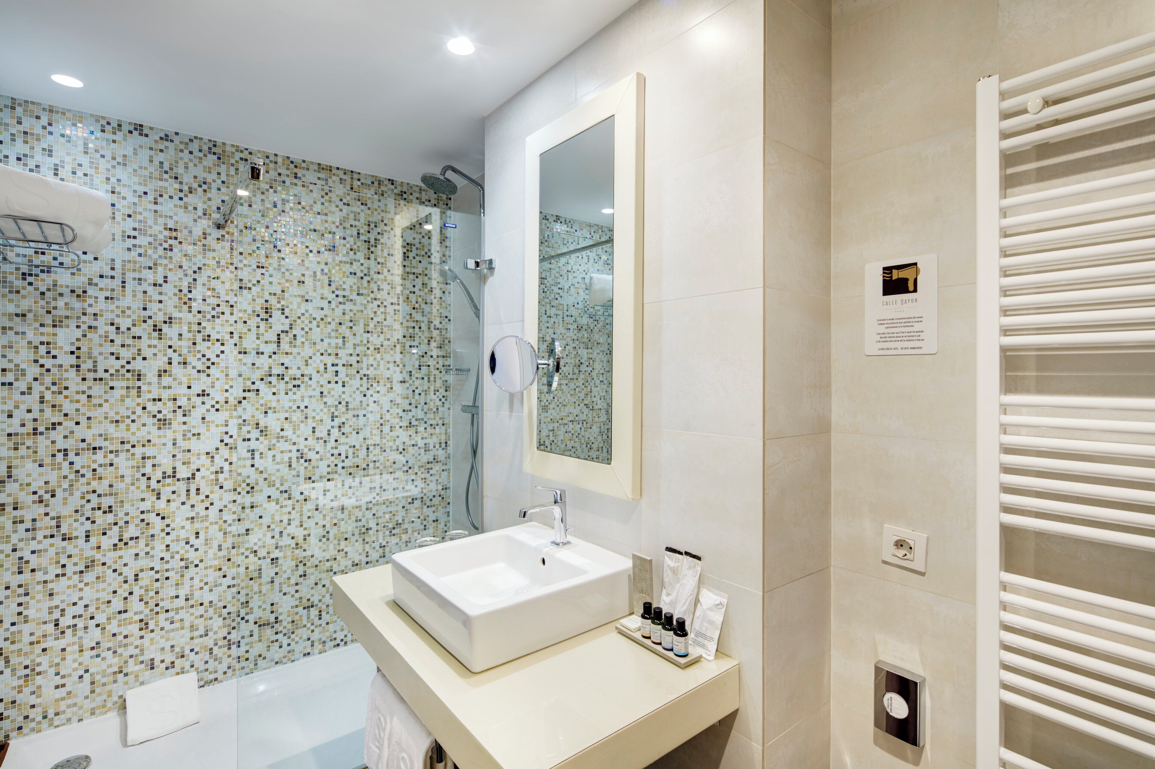 Ein modernes Badezimmer mit Duschwand aus Mosaikfliesen, einem großen Spiegel, einem weißen Waschbecken und einem Handtuchhalter.