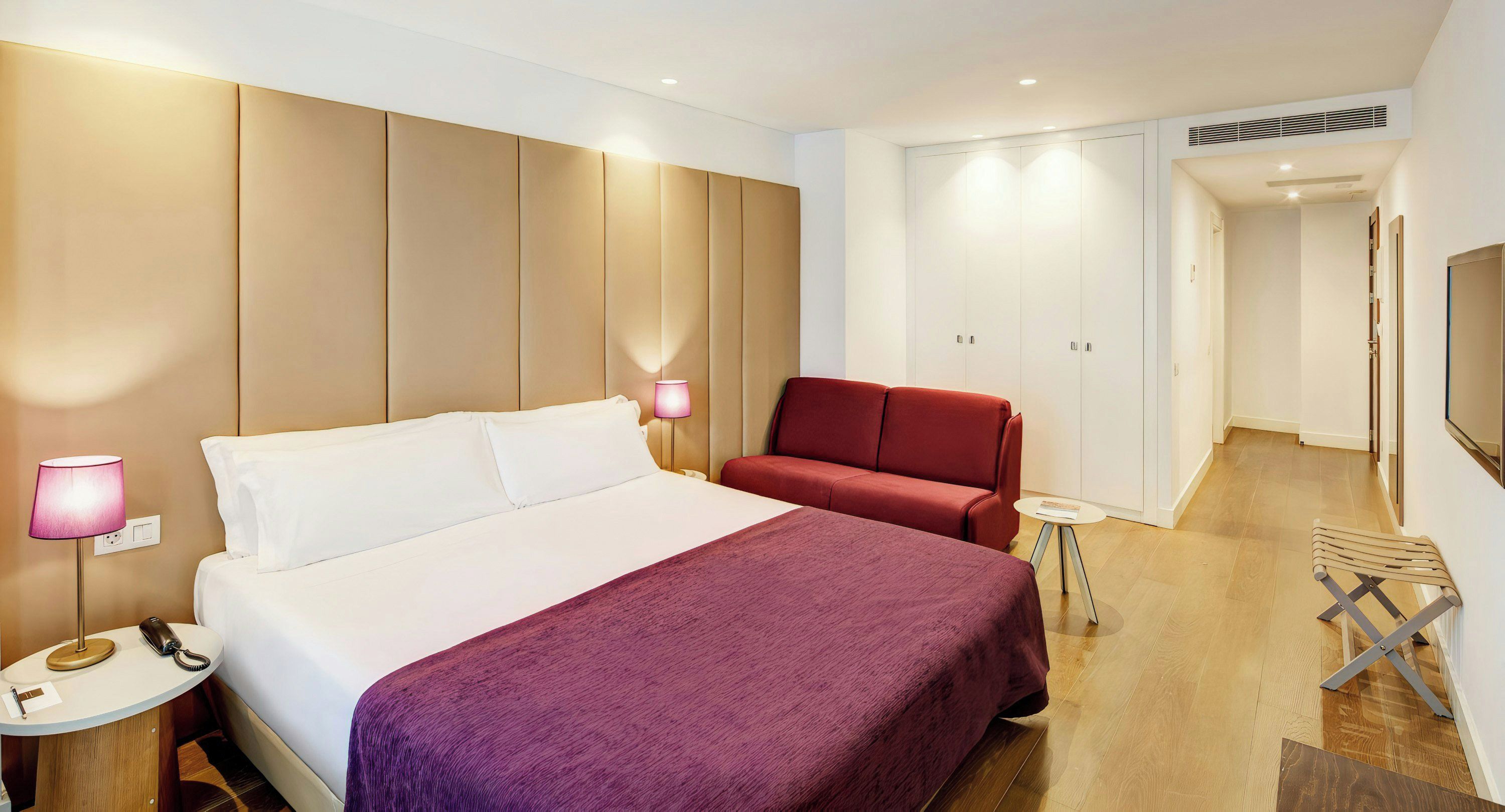Hotellihuone, jossa on sänky, punainen sohva, pieni pöytä, lamppu ja televisio.