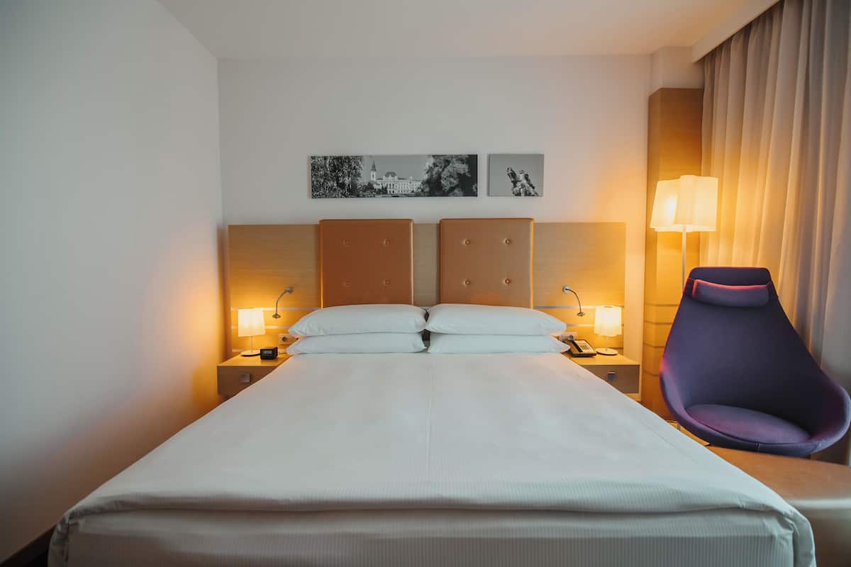 Habitación de hotel con una cama grande, dos mesitas de noche con lámparas, una silla y dos cuadros colgados en la pared.