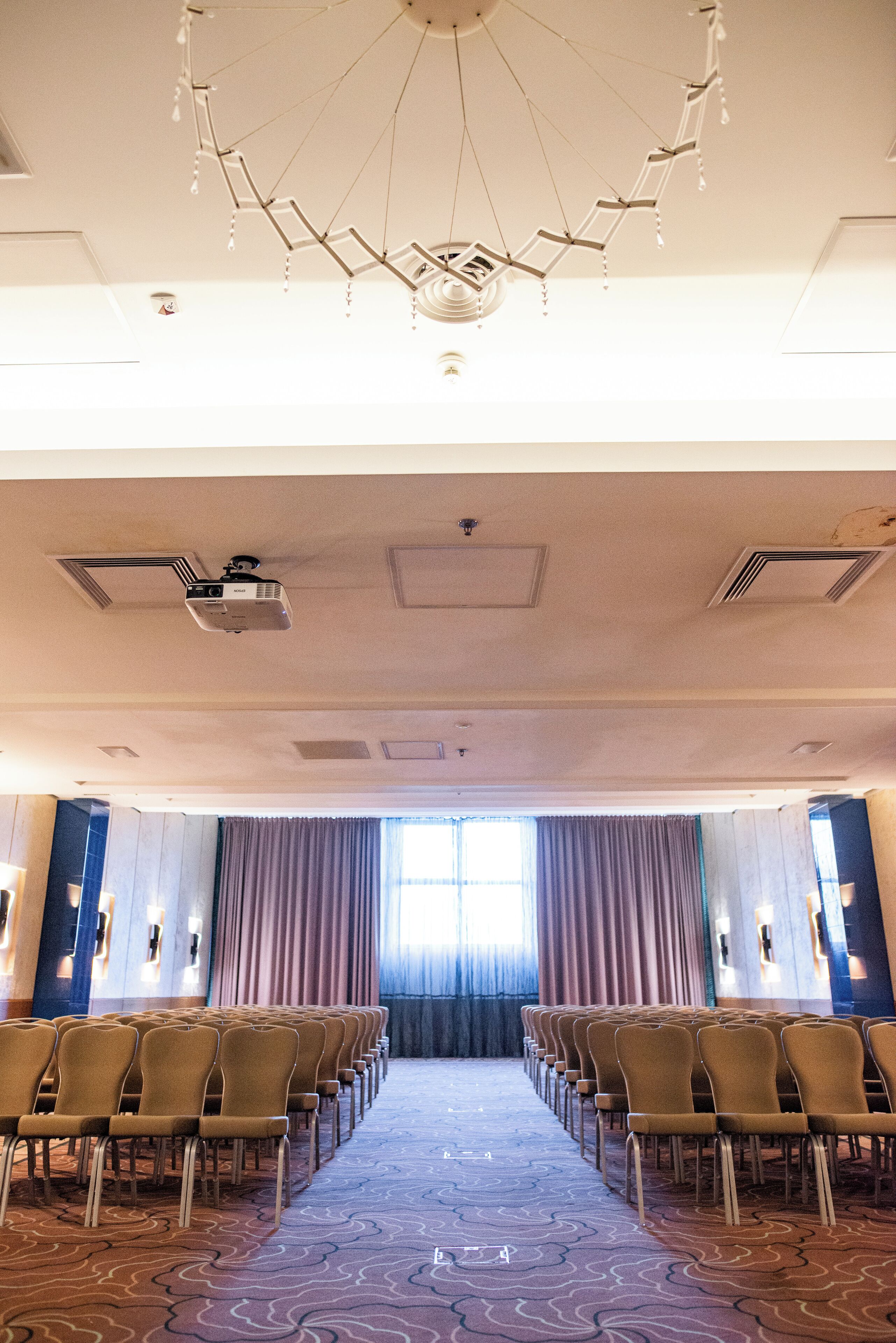 Un salón de conferencias con filas de sillas, un proyector y un amplio ventanal con cortinas.