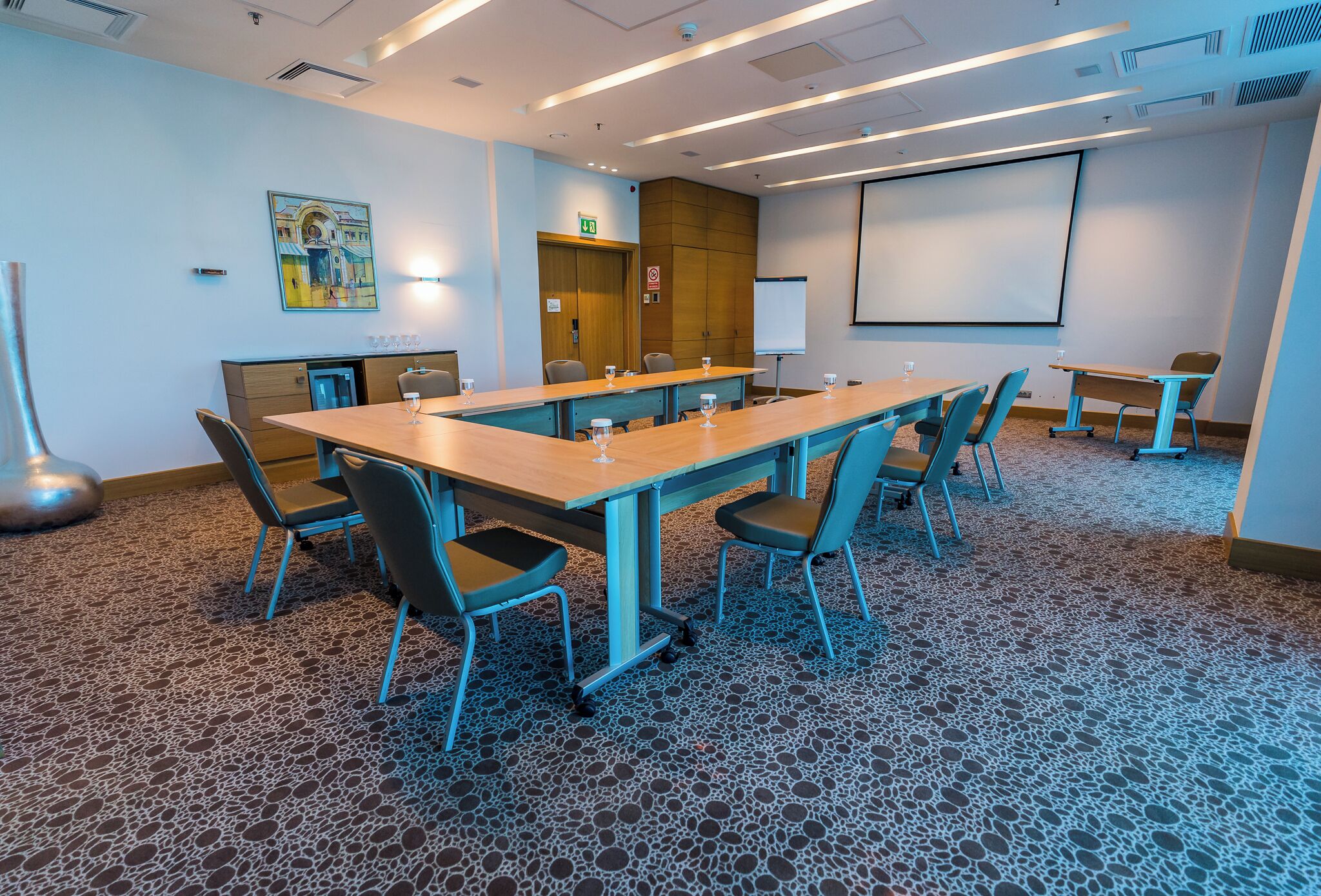 Un salón de conferencias con una larga mesa, sillas, una pantalla de proyector y una pantalla pequeña.