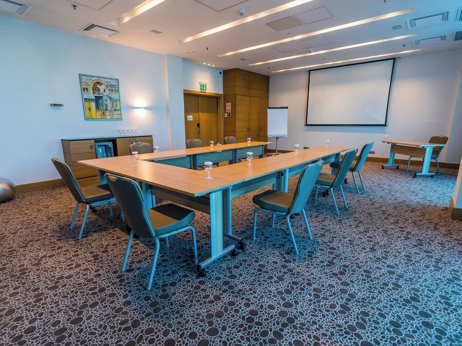Un salón de conferencias con una larga mesa, sillas, una pantalla de proyector y una pantalla pequeña.