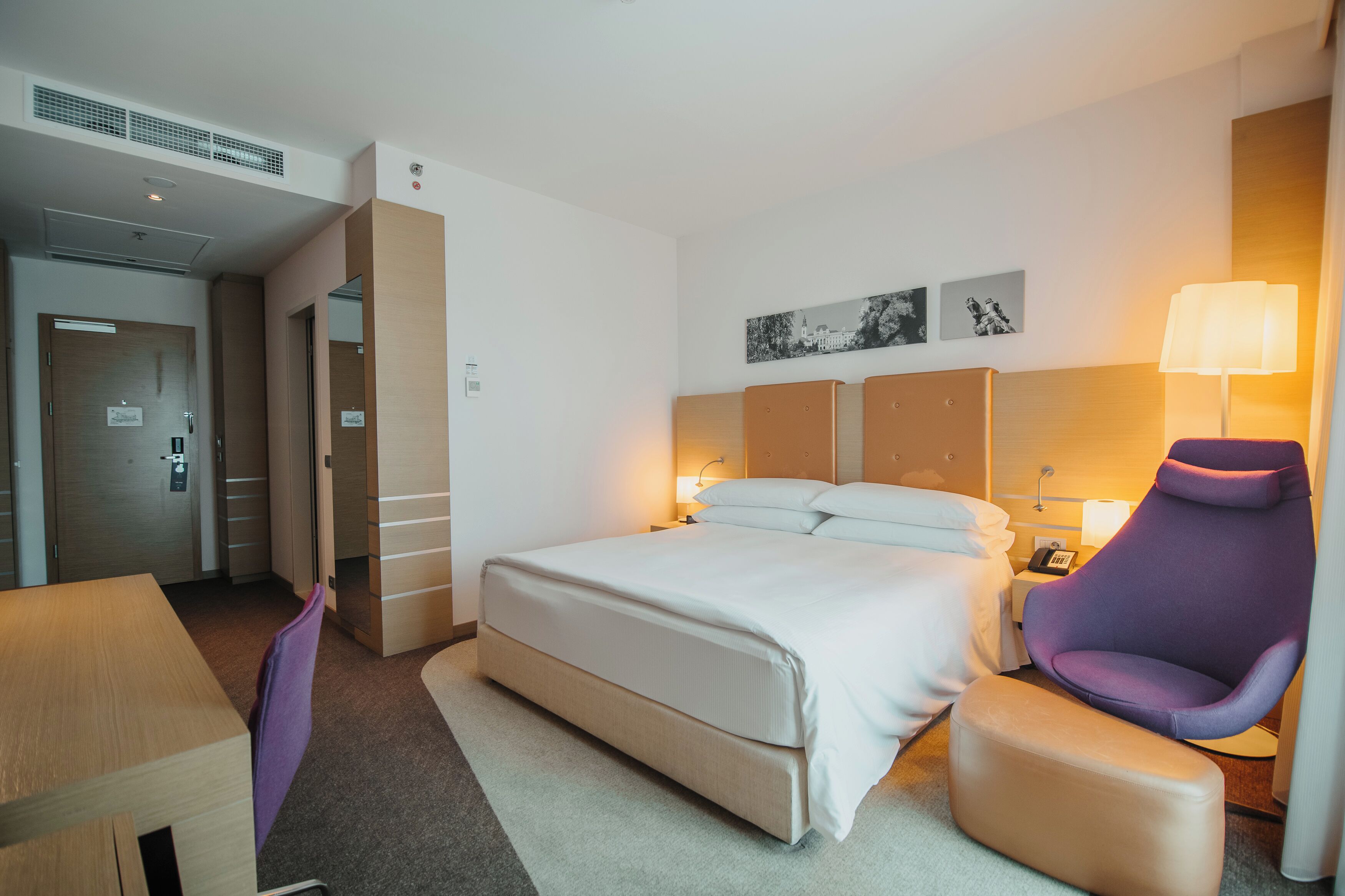 Ett hotellrum med en säng, en stol, ett skrivbord och ett nattduksbord med en lampa.