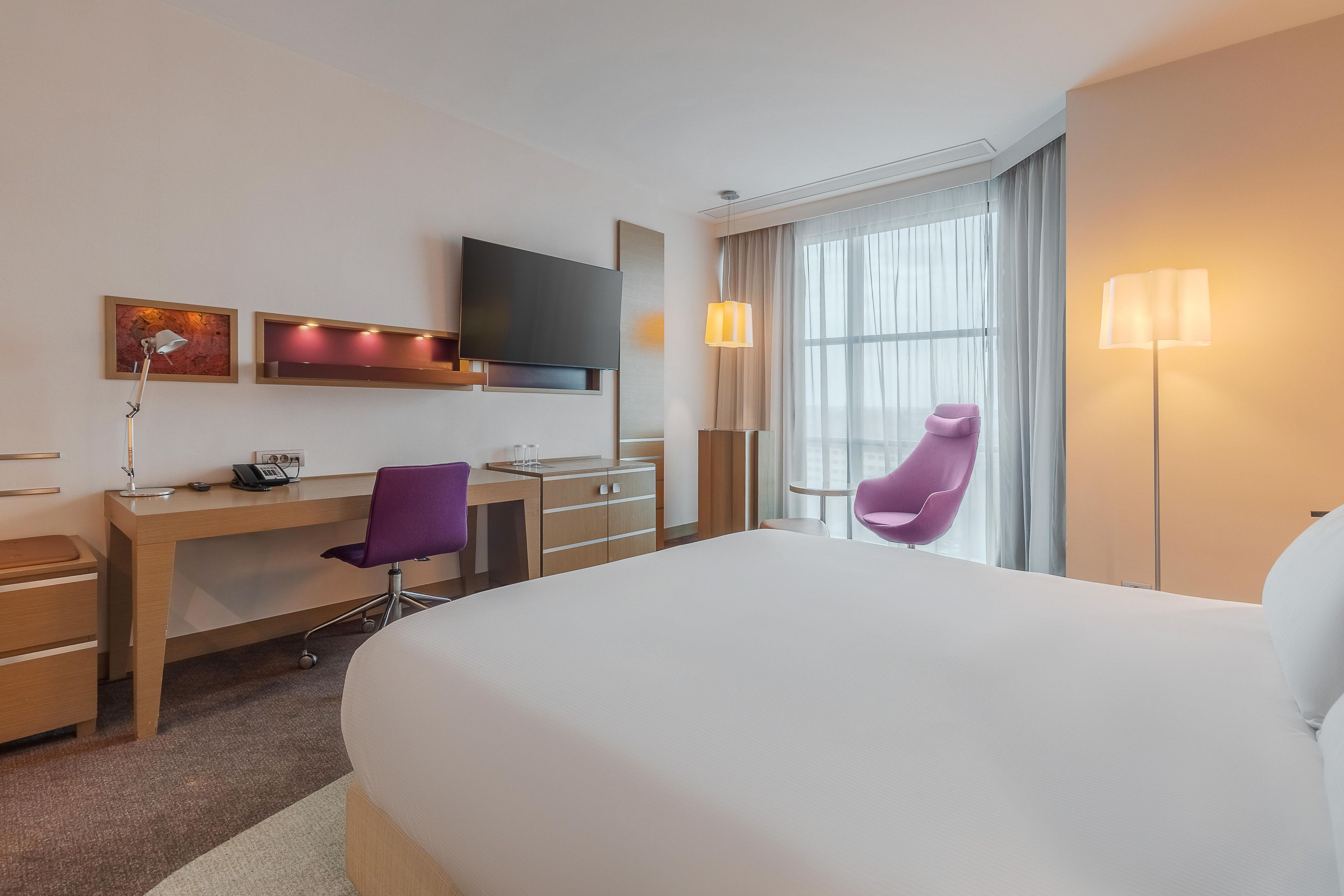 Habitación de hotel con una cama grande, un escritorio con silla, un televisor de pantalla plana y una ventana con cortinas.