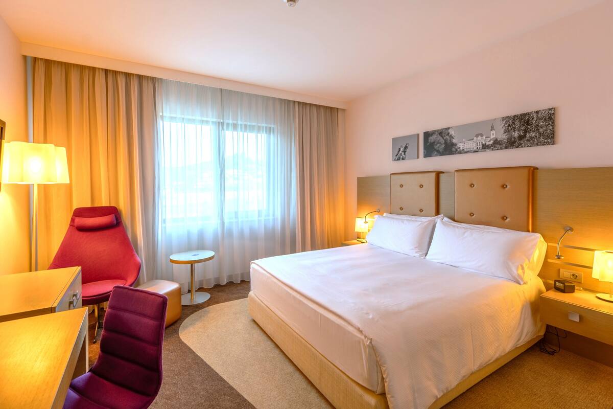 Habitación de hotel con una cama grande, un escritorio, una silla, una mesita y una ventana con cortinas.