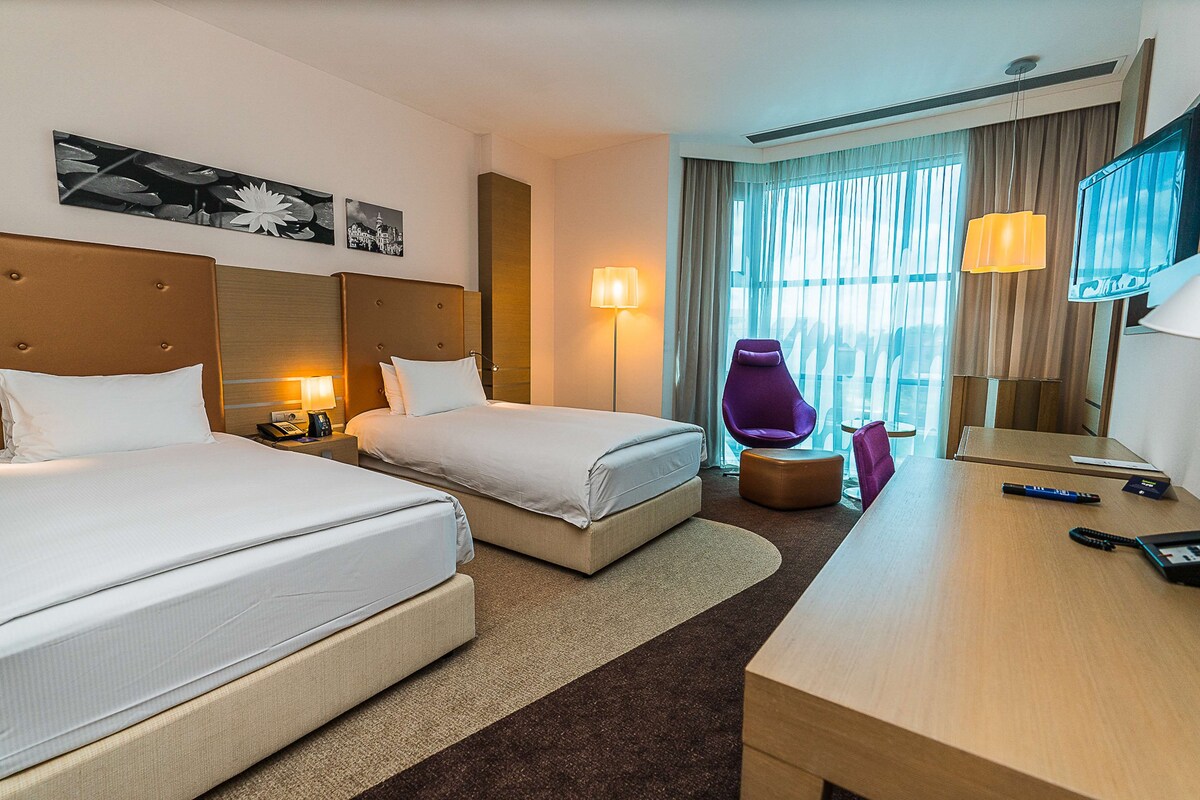 A hotel room with two beds, a desk, a chair, a TV, and a window with curtains.