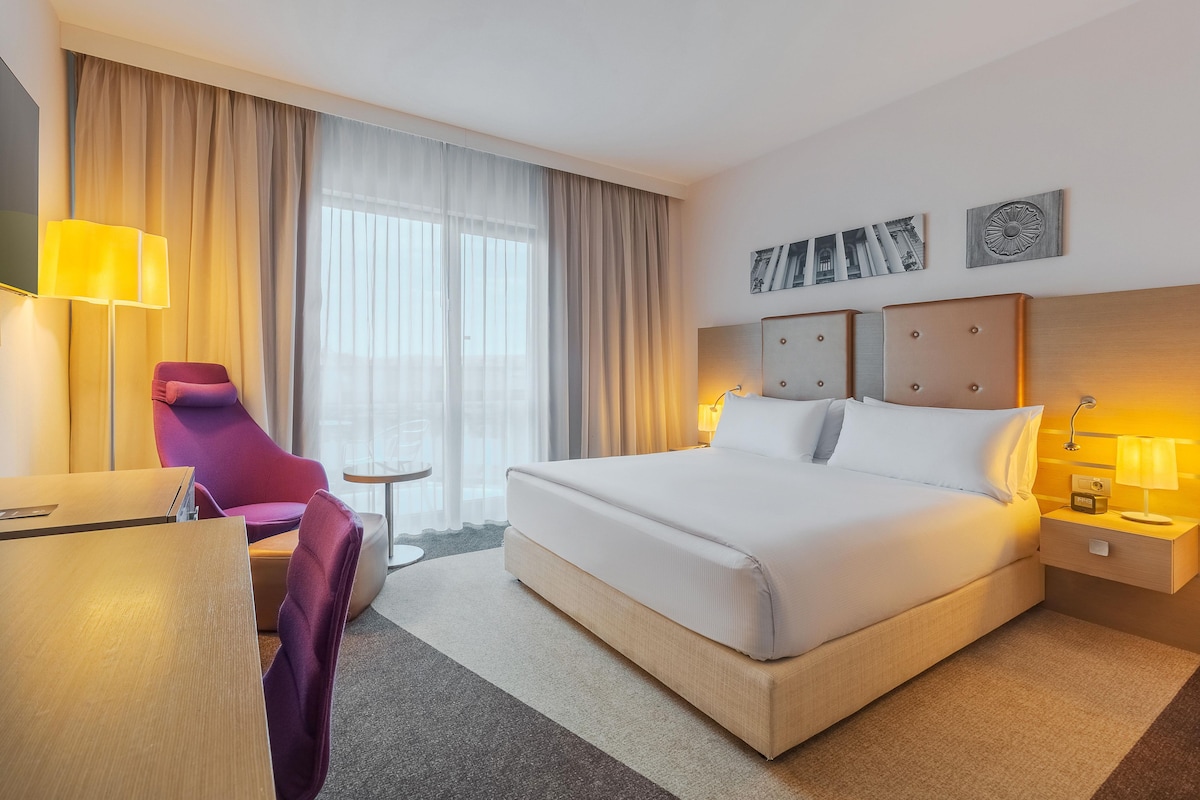 Une chambre d’hôtel avec un grand lit, un bureau avec une chaise, une petite table et une fenêtre avec des rideaux.