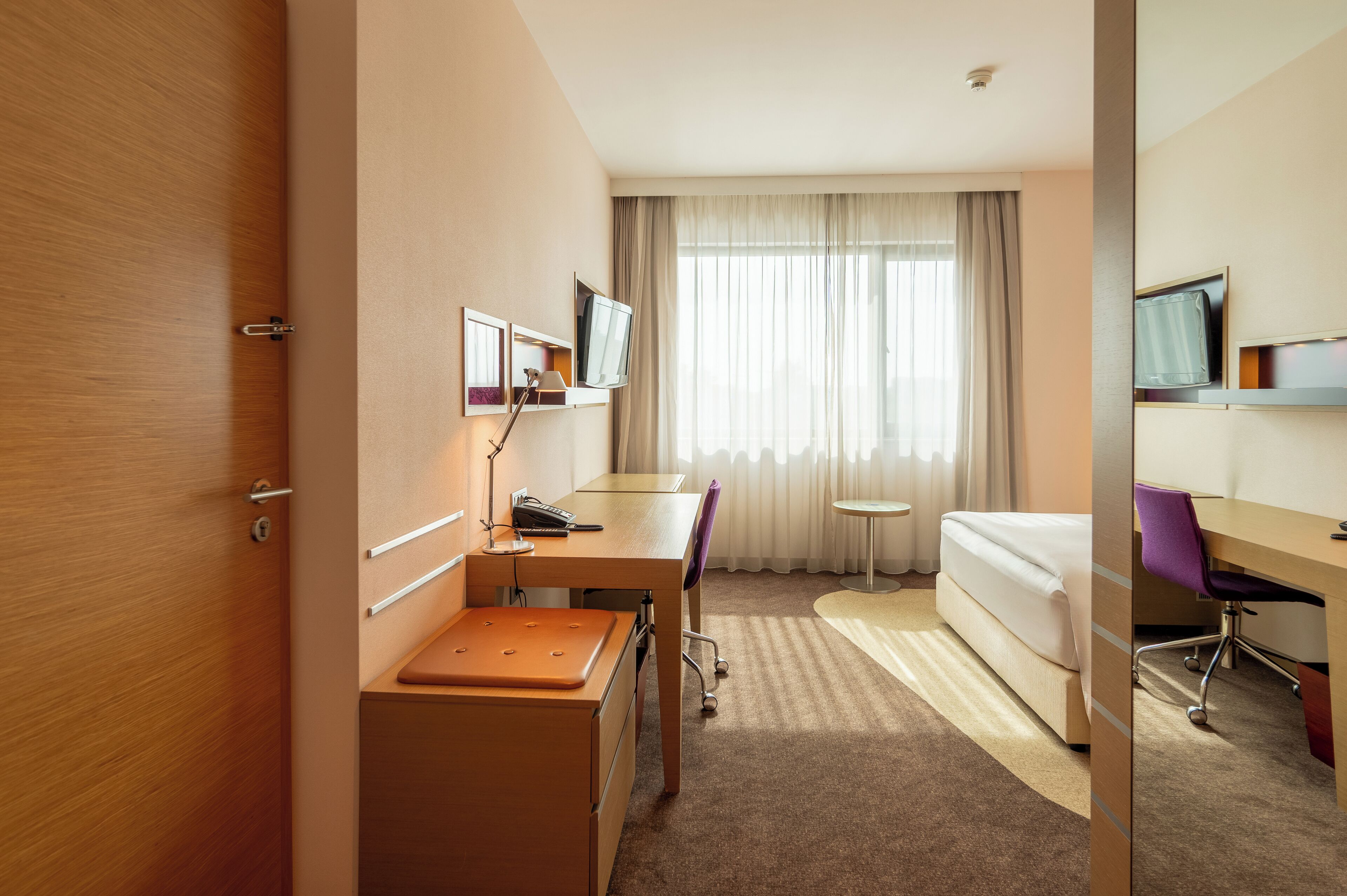 Ein Hotelzimmer mit Holztür, Schreibtisch mit Stuhl, Bett, kleinem Tisch und Fernseher an der Wand.