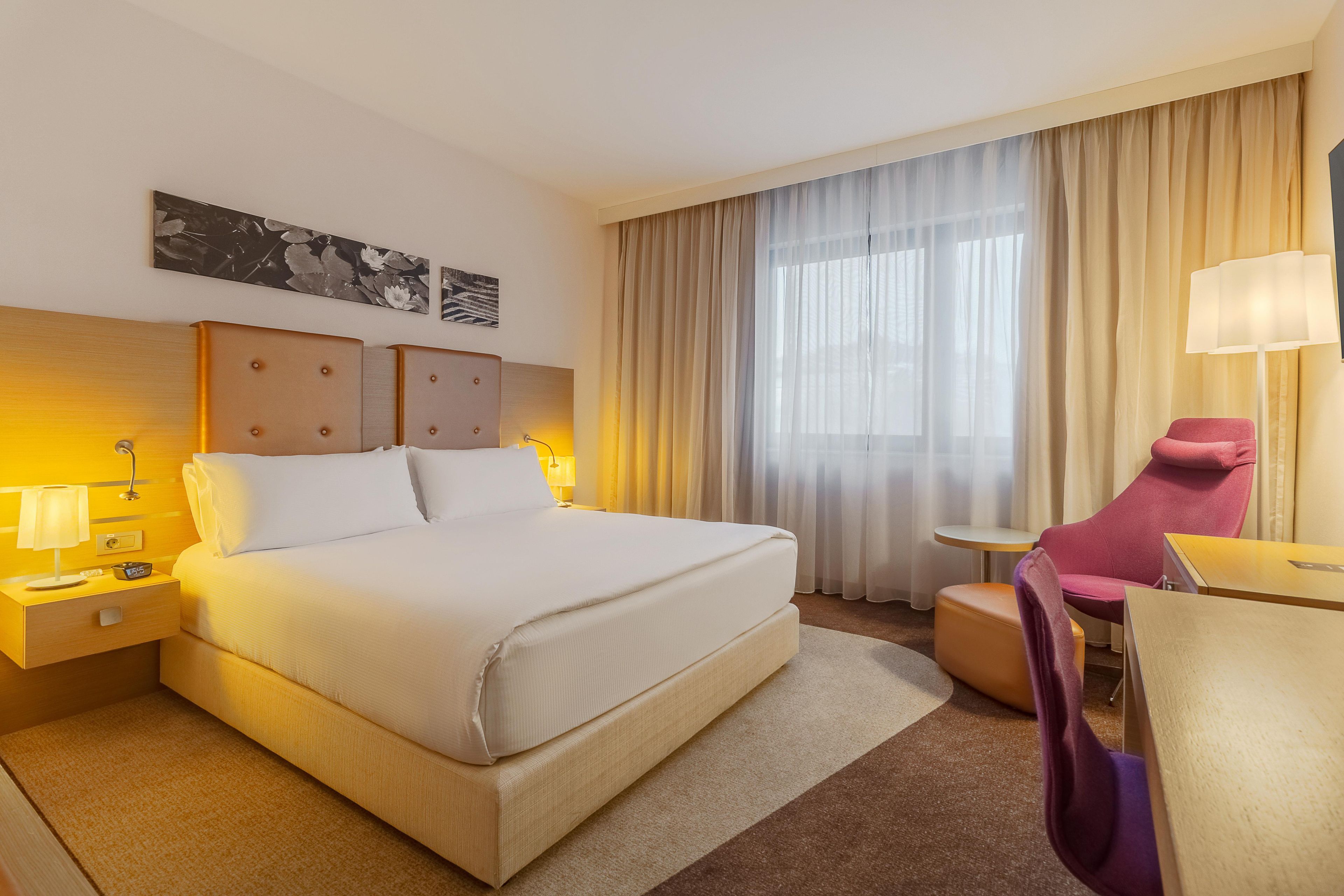 Habitación de hotel con una cama grande, un escritorio, una silla, una mesita de noche, una lámpara y una ventana con cortinas.