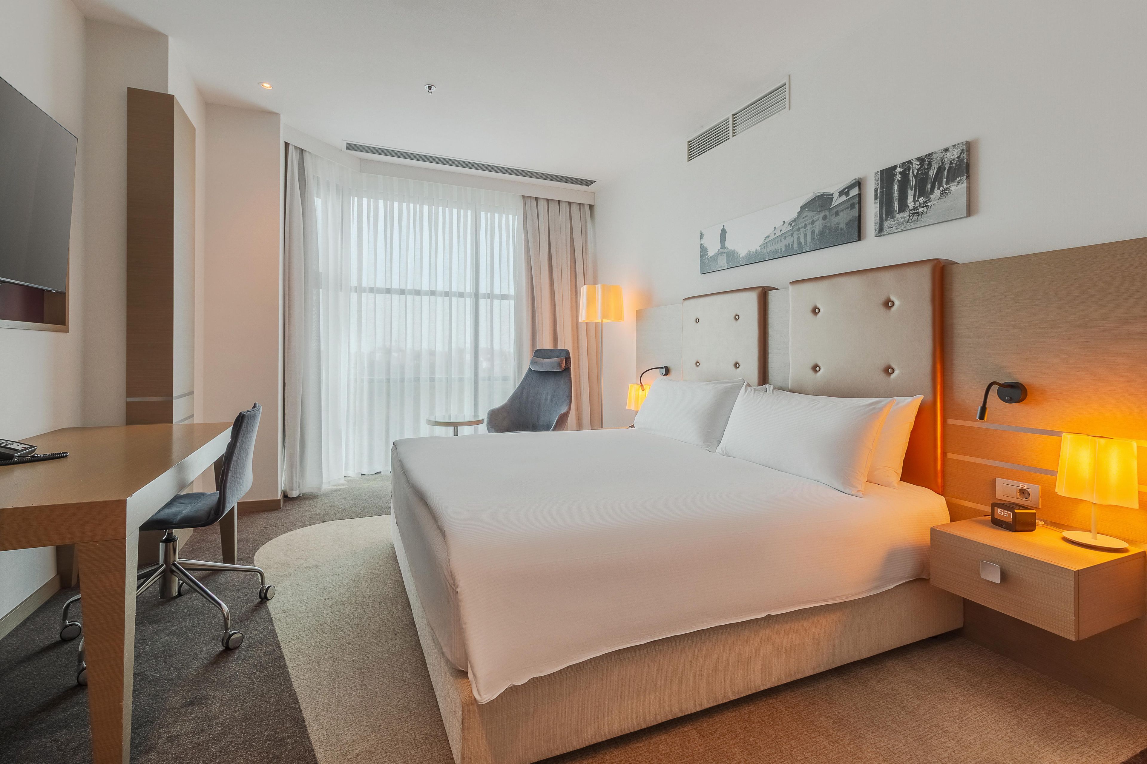 A hotel room with a bed, bedside table, lamp, chair, desk, and a television.