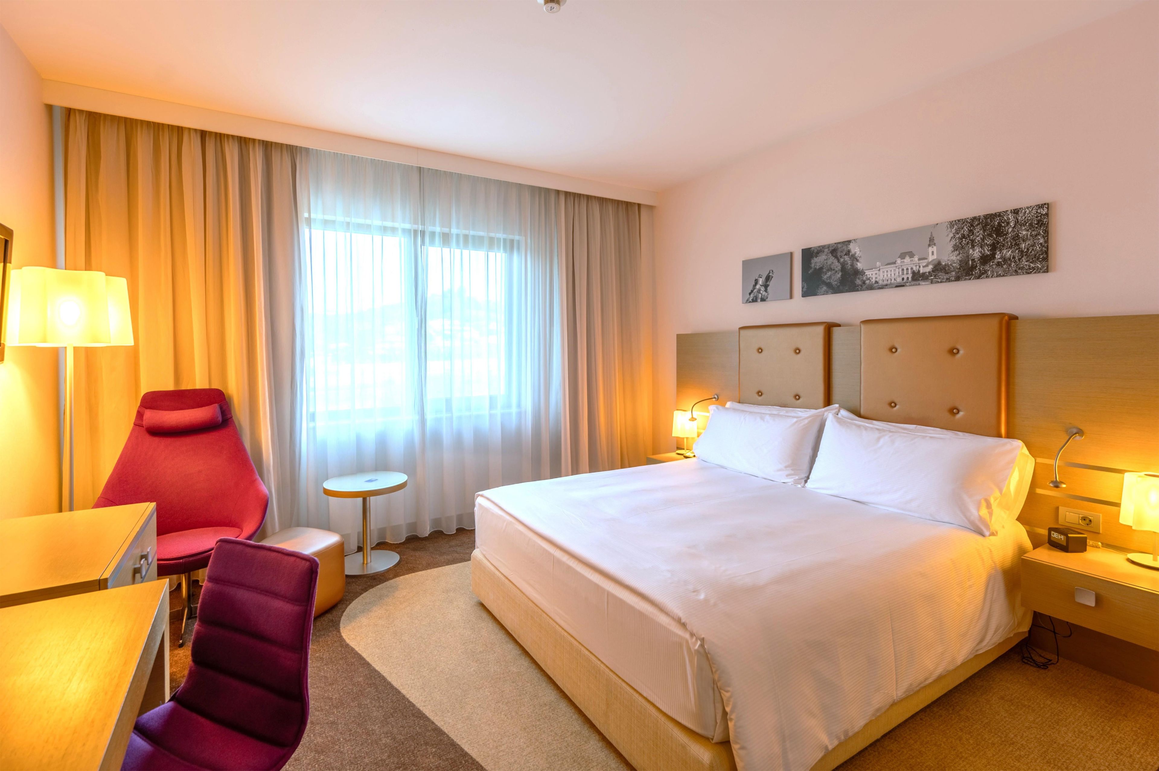 Een hotelkamer met een groot bed, een bureau, een stoel, een klein tafeltje en een raam met gordijnen.