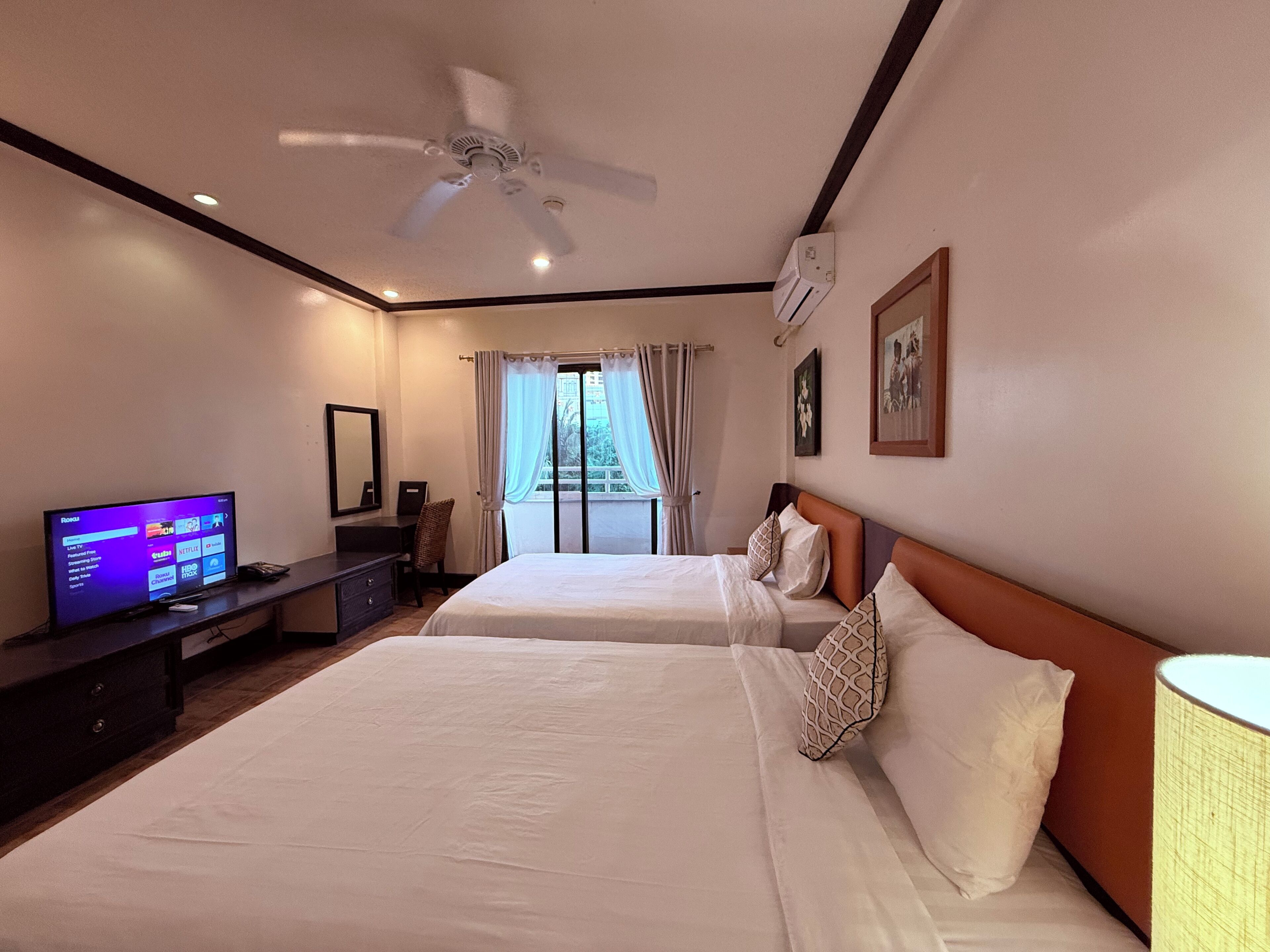 A hotel room with two beds, a TV, a ceiling fan, and a window with curtains.