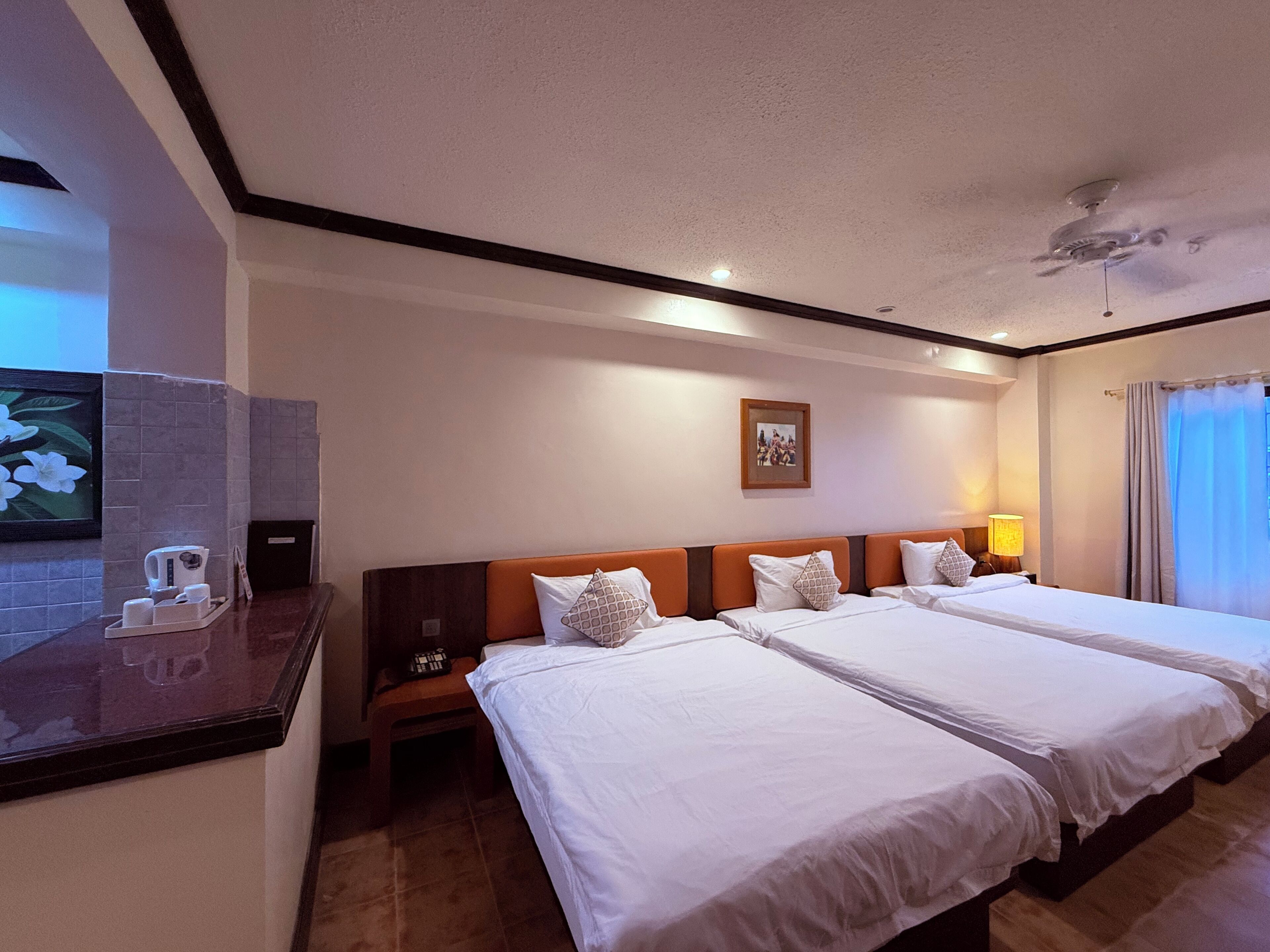 A hotel room with two beds, a small table with a kettle and cups, a nightstand, a framed picture on the wall, and a window with curtains.