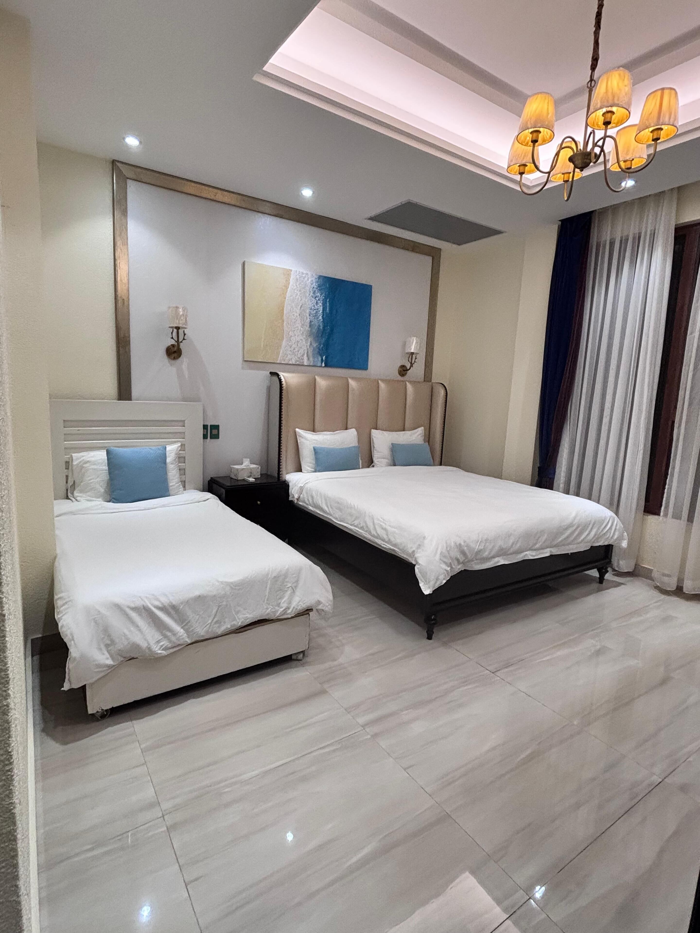A modern bedroom with two beds, a large painting, and a chandelier.