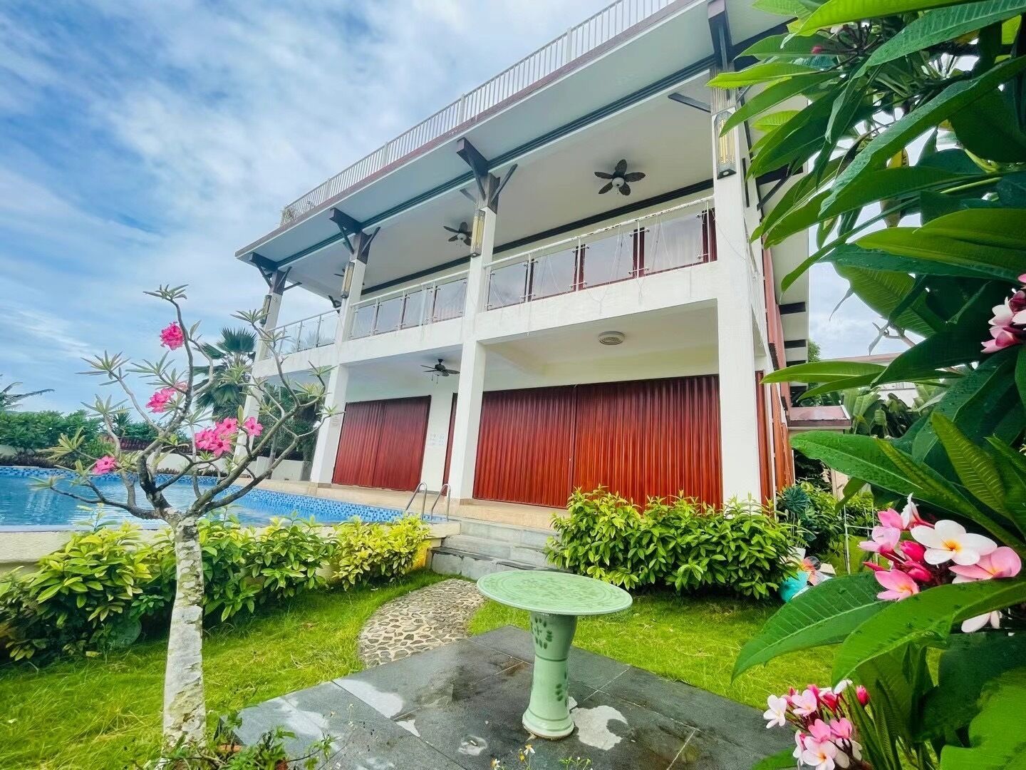 A two-story building with a pool, surrounded by lush greenery and flowering plants.