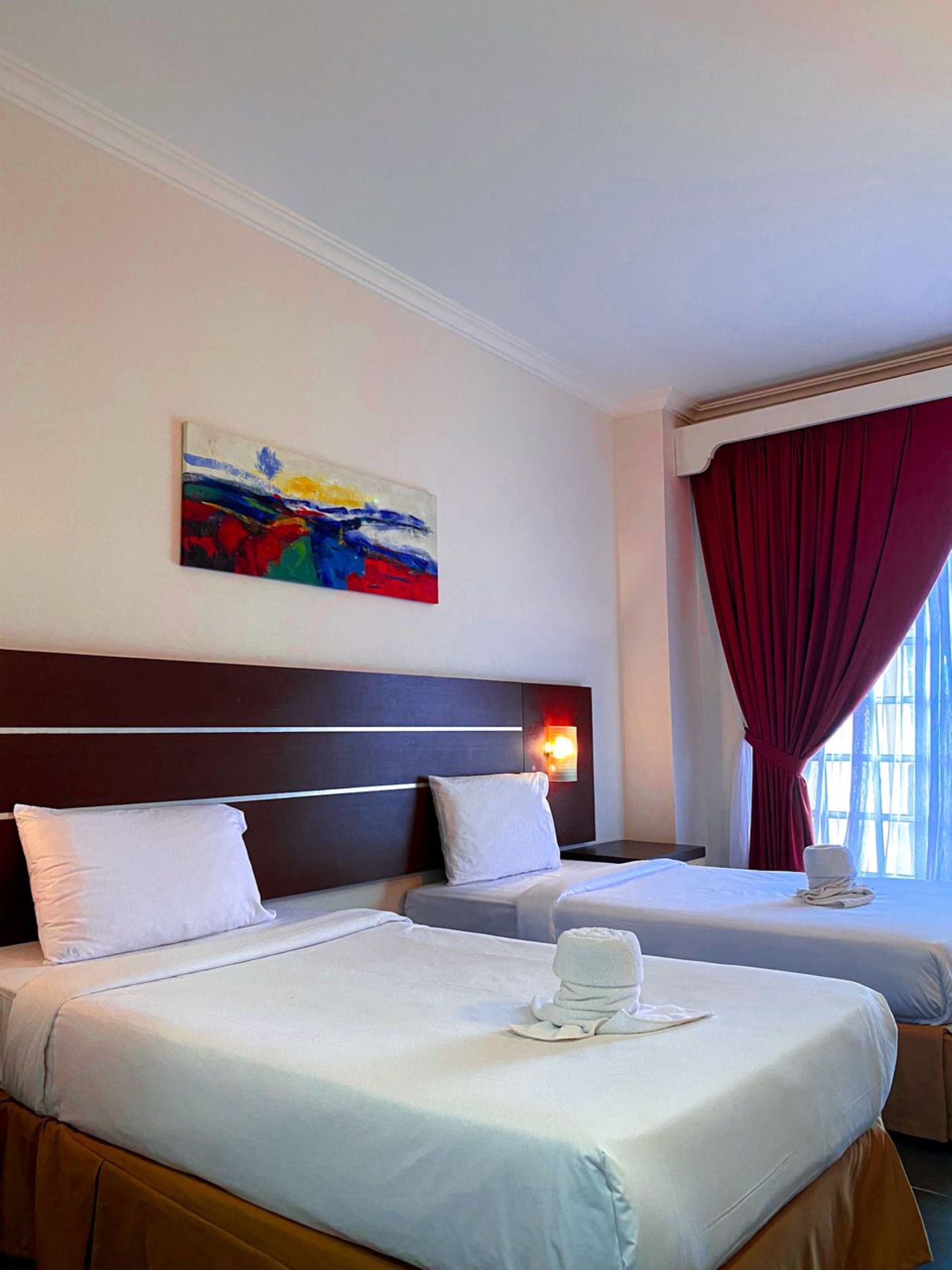 A hotel room with two beds, a painting on the wall, a night light, and a window with red curtains.