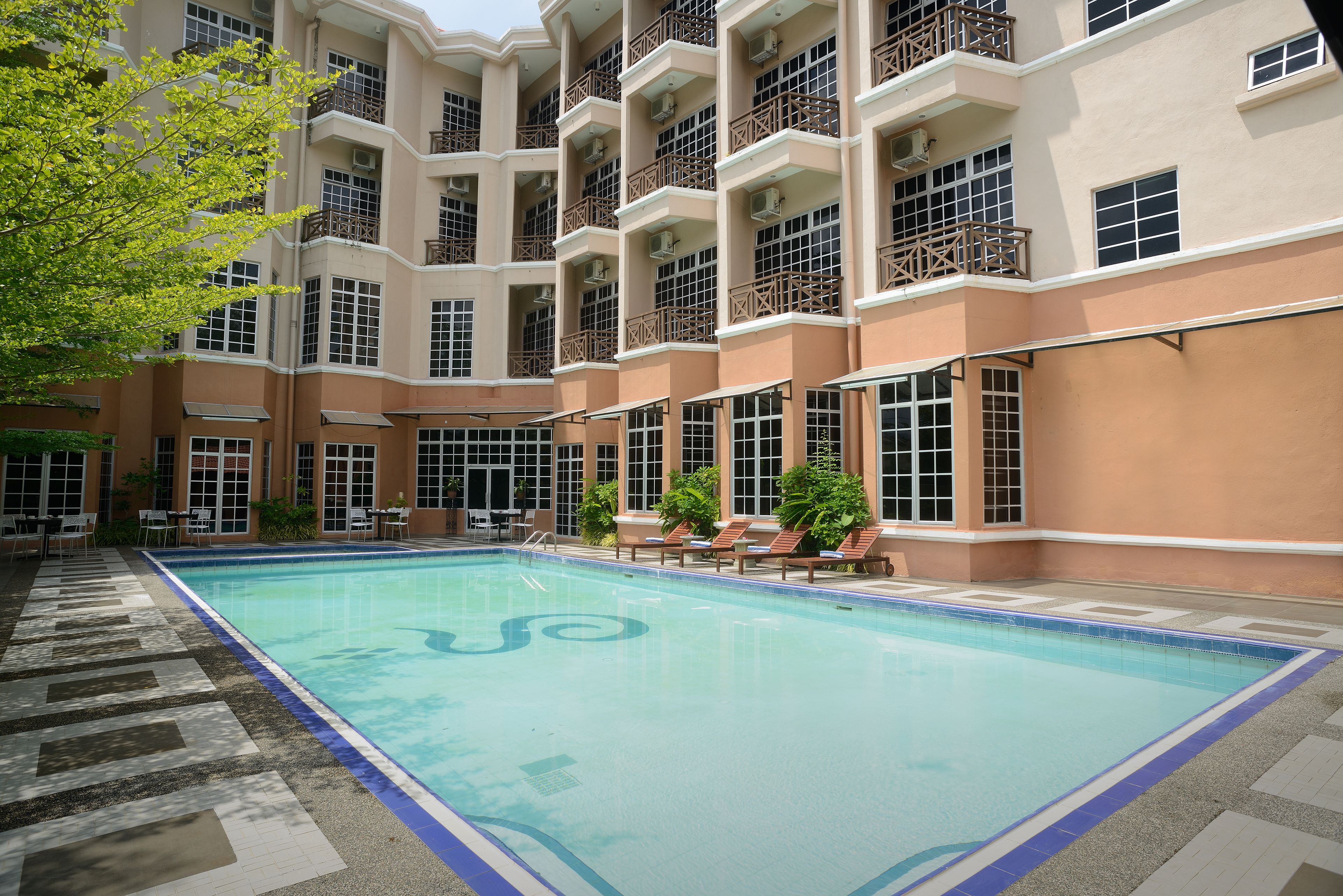 A swimming pool with a tiled deck, surrounded by a multi-story building with balconies and windows.