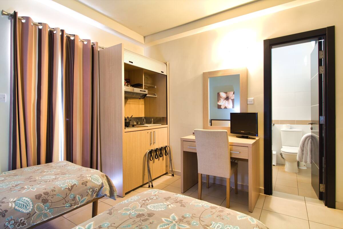 Quarto de hotel com duas camas, uma escrivaninha, uma cadeira, uma televisão, banheiro e armário.