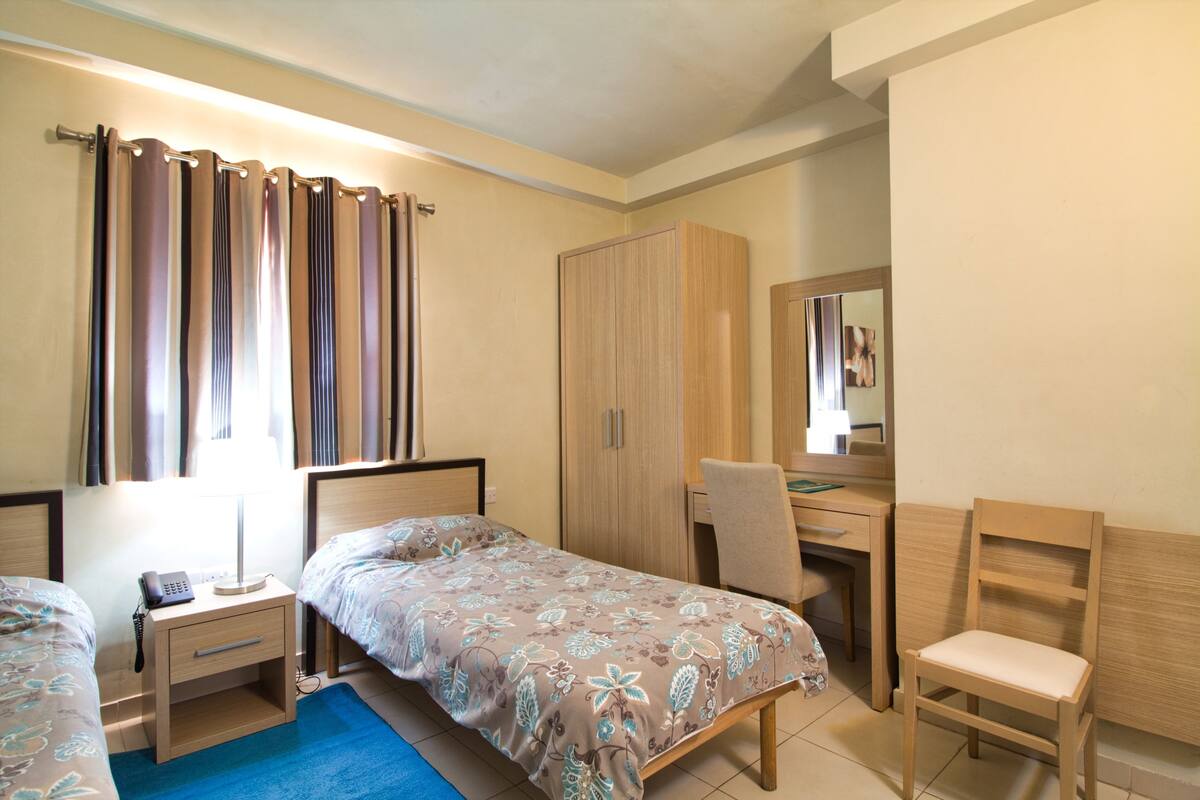 Quarto com duas camas de solteiro, uma escrivaninha, uma cadeira, um guarda-roupa e uma mesa de cabeceira com abajur.